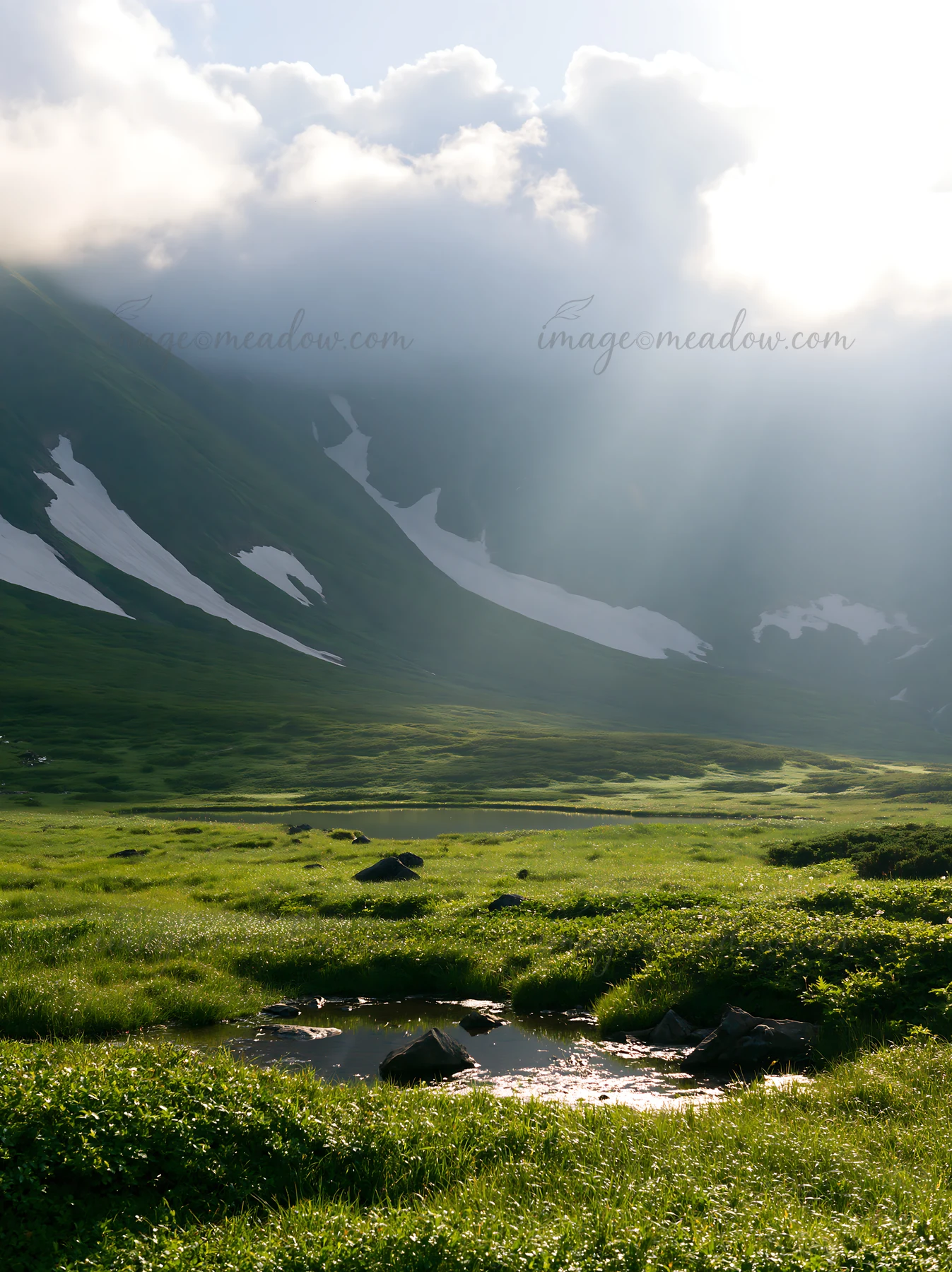 Light rays break through clouds over the Daisetsuzan alpine meadow. Green grass covers the landscape, with snow patches on the mountains. A small pond reflects