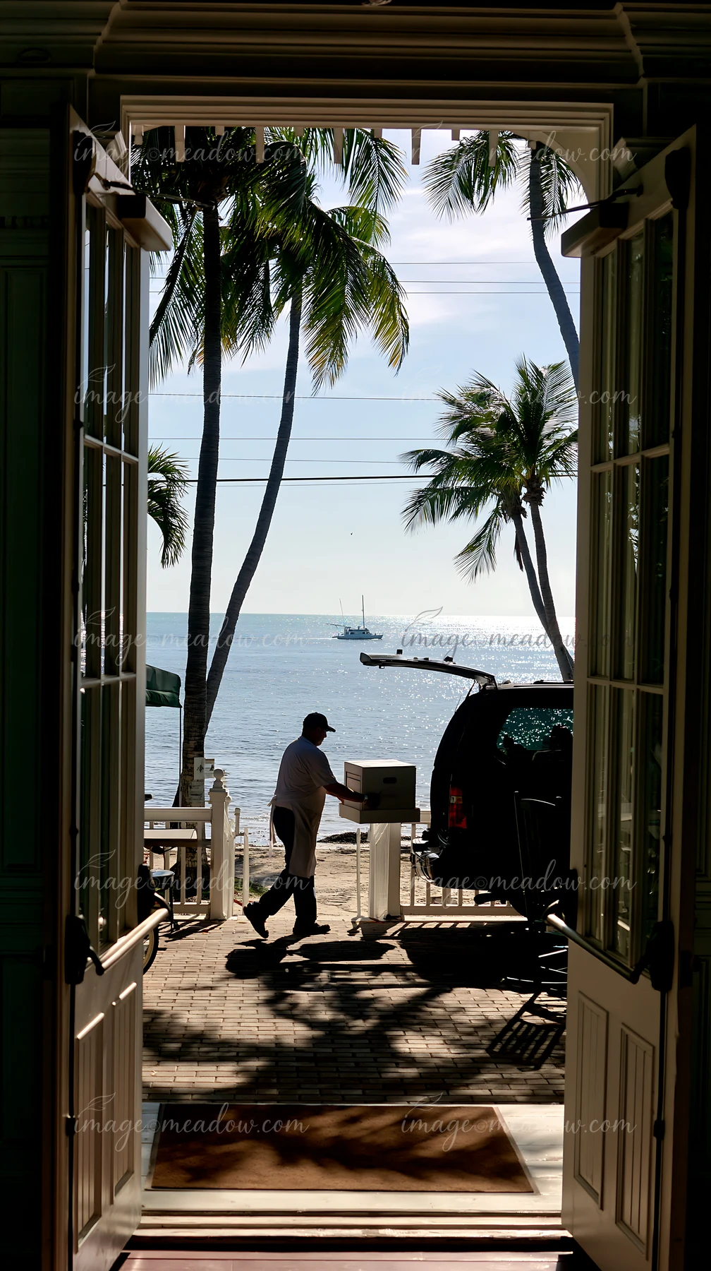 Delivery worker carries a cardboard box toward a seaside venue through open doors, with palm trees and ocean beyond.