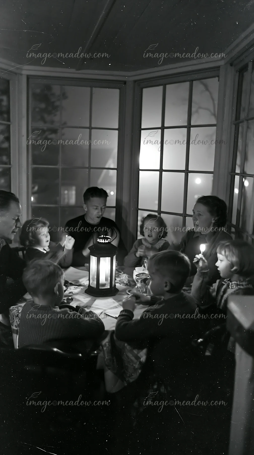 Family sings around a glowing lantern at a dining table indoors near large windows.