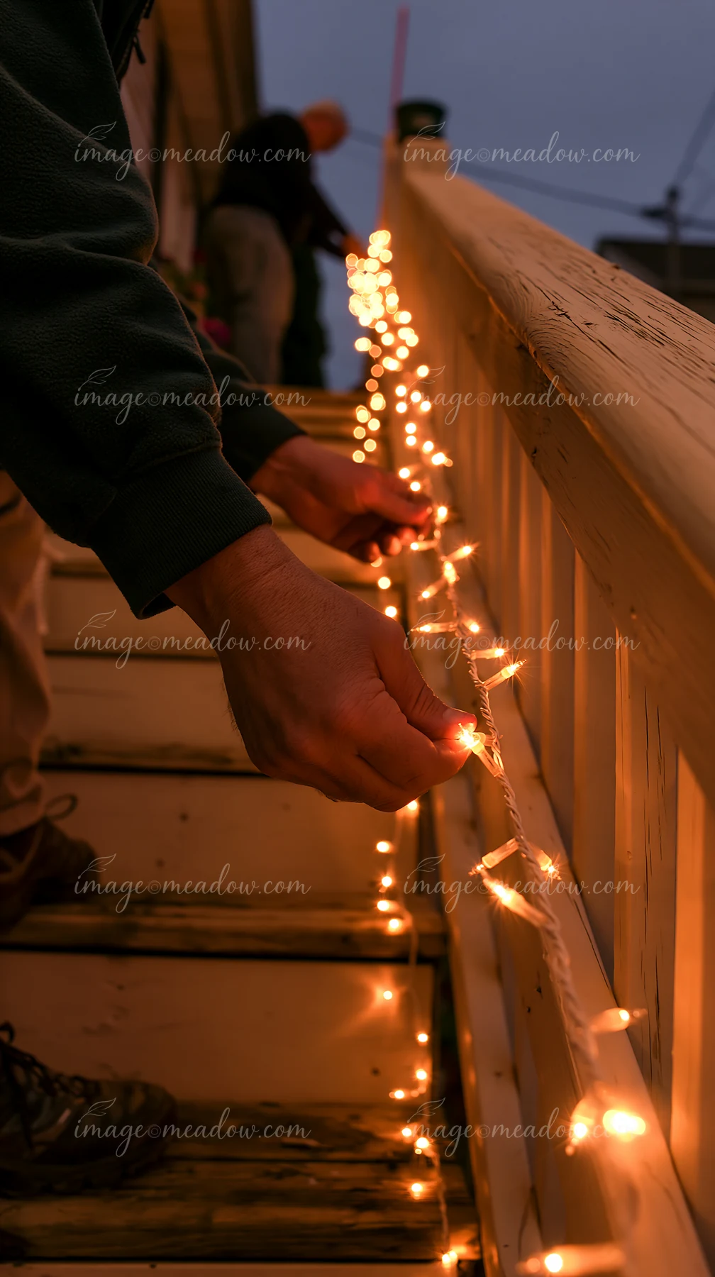 Hands attach glowing string lights to a wooden staircase railing in a dim interior.