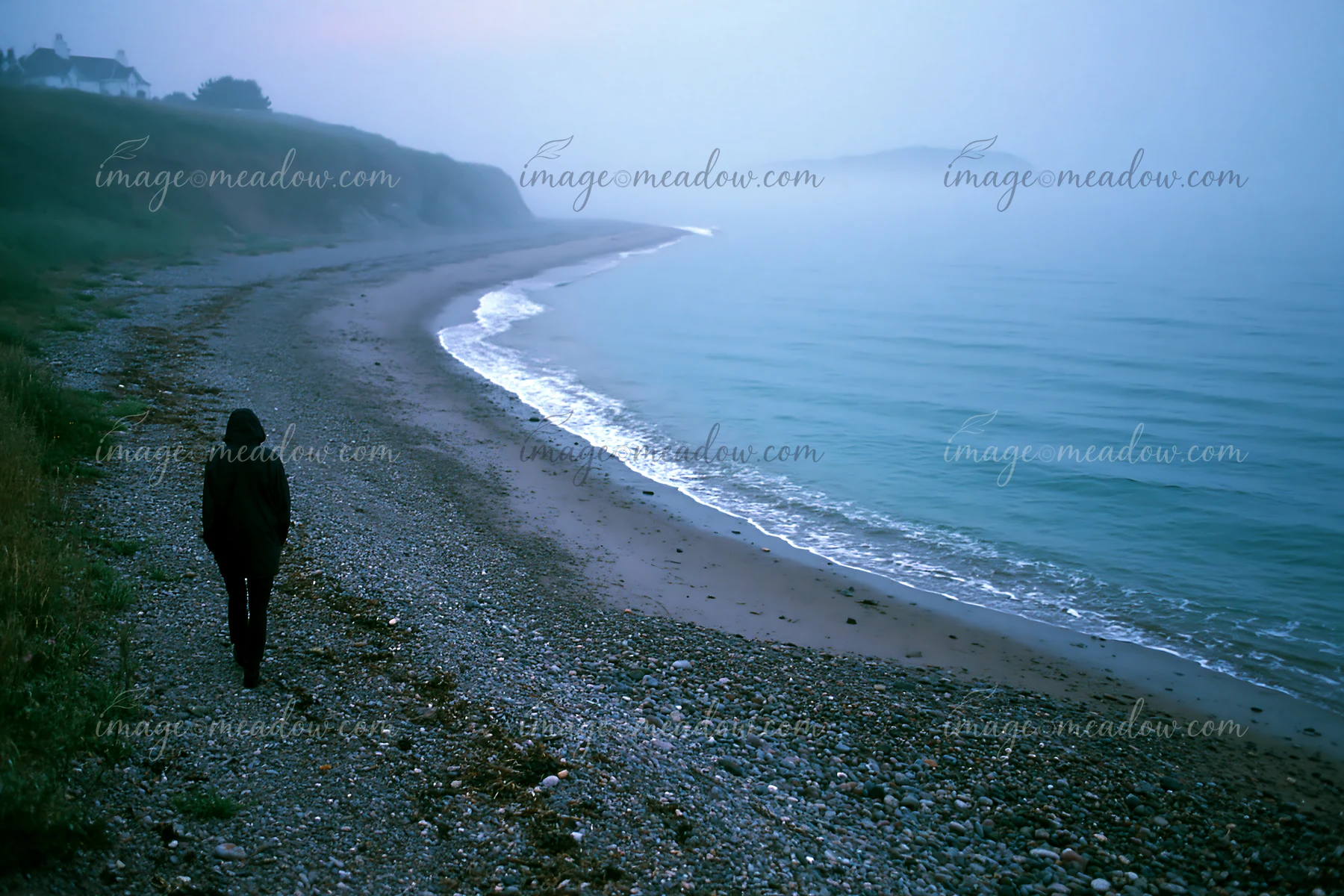 Hooded person walks alone along a misty pebble beach beside calm coastal water and cliffs.