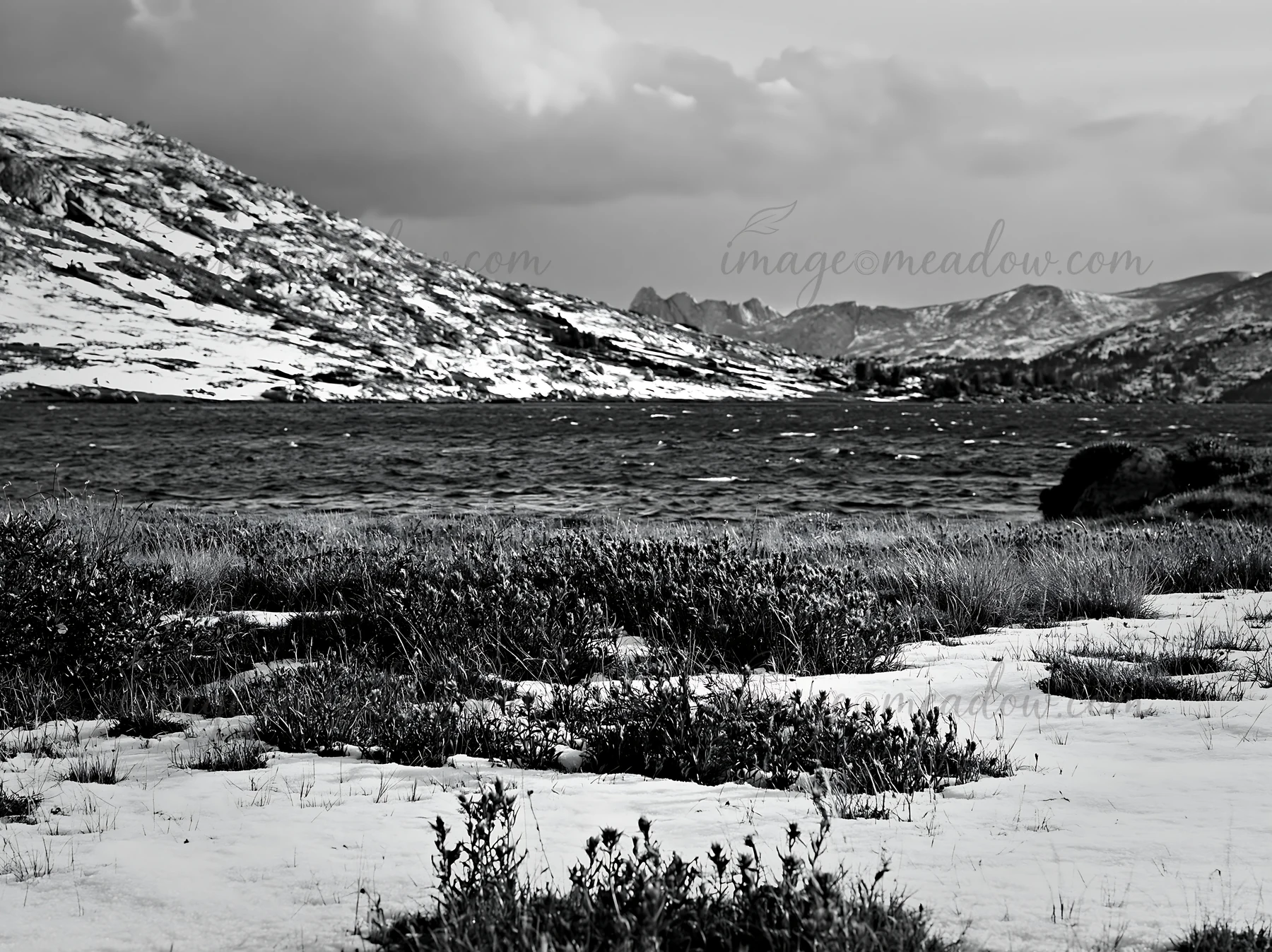 A lakeside meadow shows snow covering the ground with patches of red plants. The scene presents distant hills and a calm body of water beneath a cloudy sky. Pre