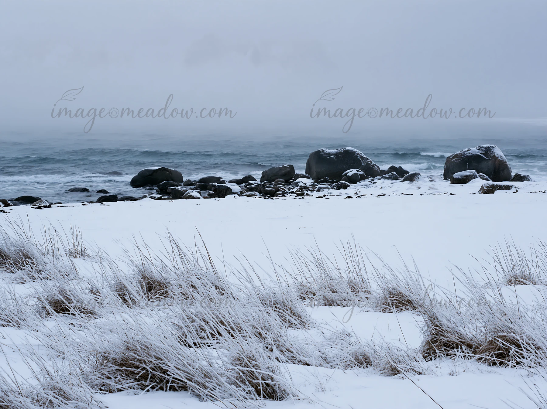 In Lofoten, a coastal meadow is blanketed with snow. Rocks emerge from the snow and contrast with the dark sea. Overcast skies create a gray atmosphere above th
