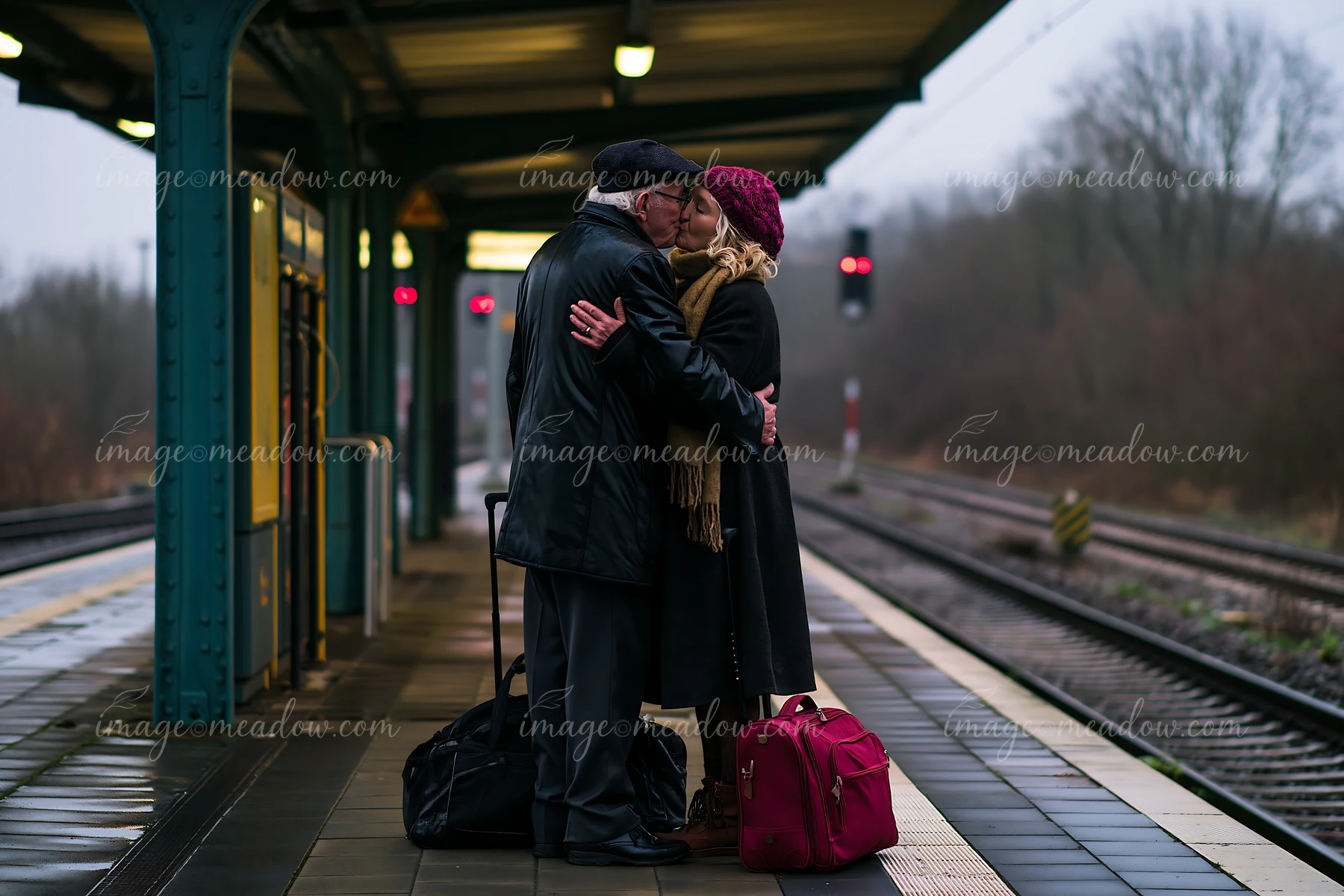 Mature couple kissing on a rainy railway platform beside their luggage.