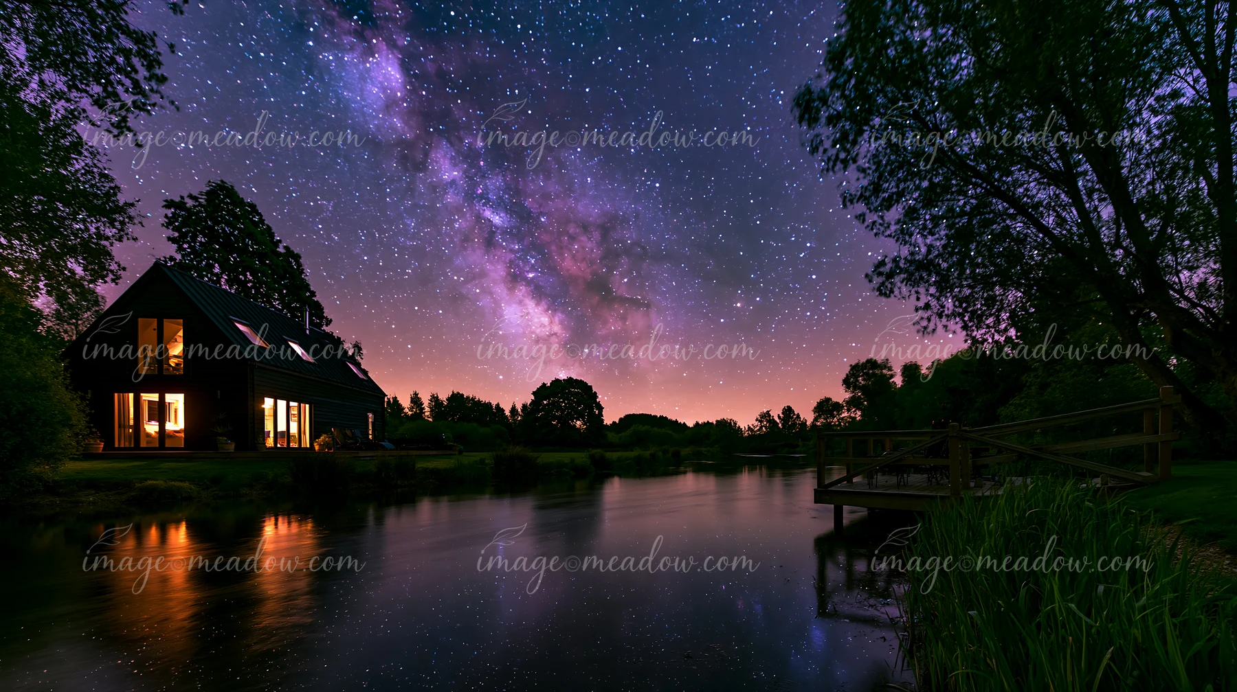 Modern house with warm windows beside a calm pond beneath the Milky Way and a star-filled night sky.