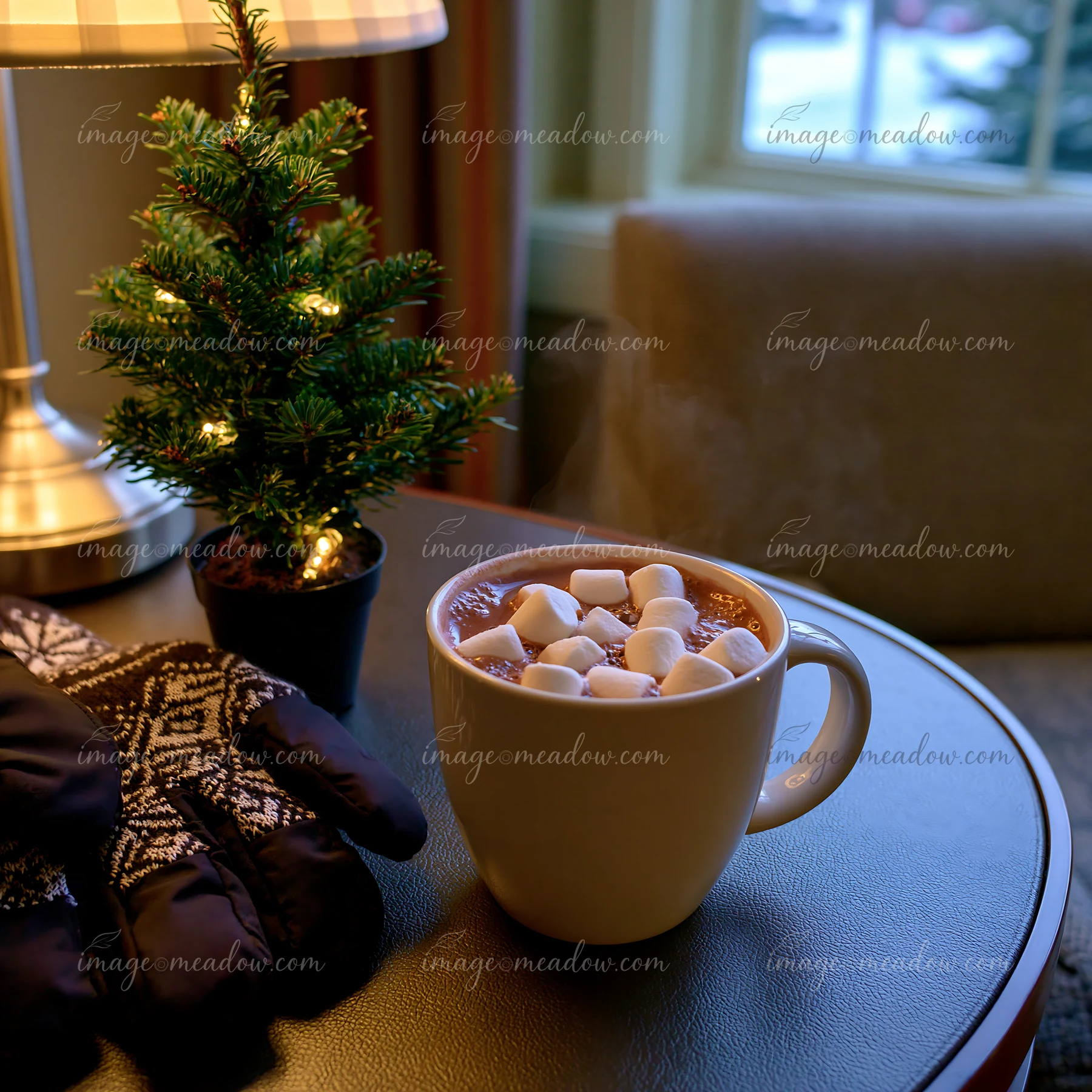 Mug of cocoa topped with marshmallows sits on a table beside a small lit evergreen tree.