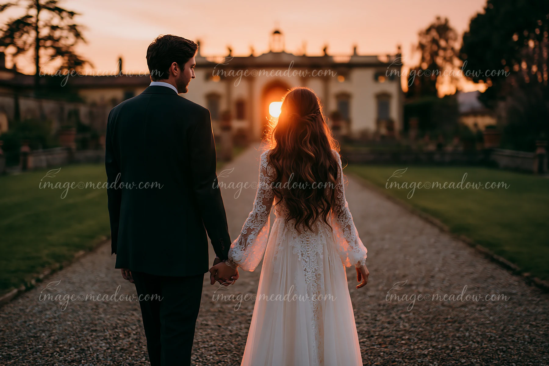 Newlywed couple holding hands as they walk through a church garden toward a building at sunset.
