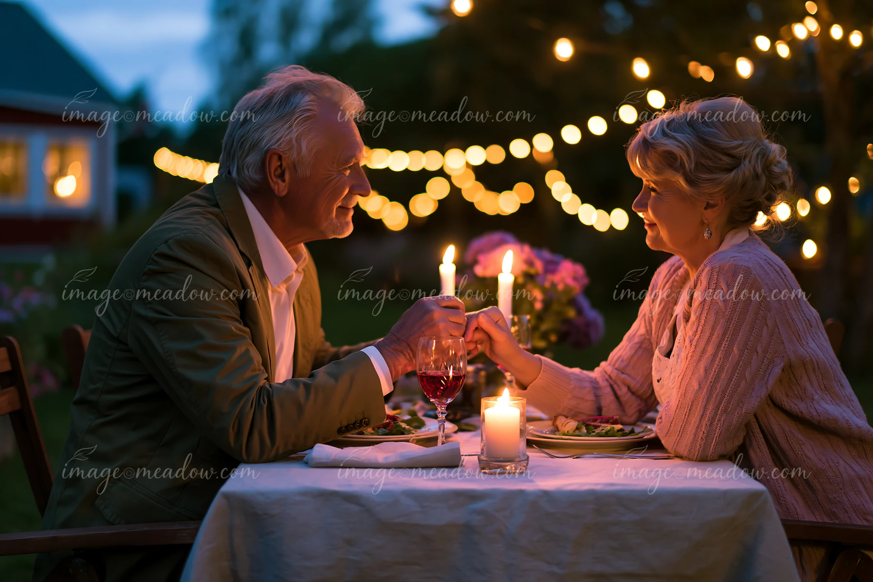 Older couple sharing a tender hand kiss at a candlelit outdoor dinner with string lights and flowers.