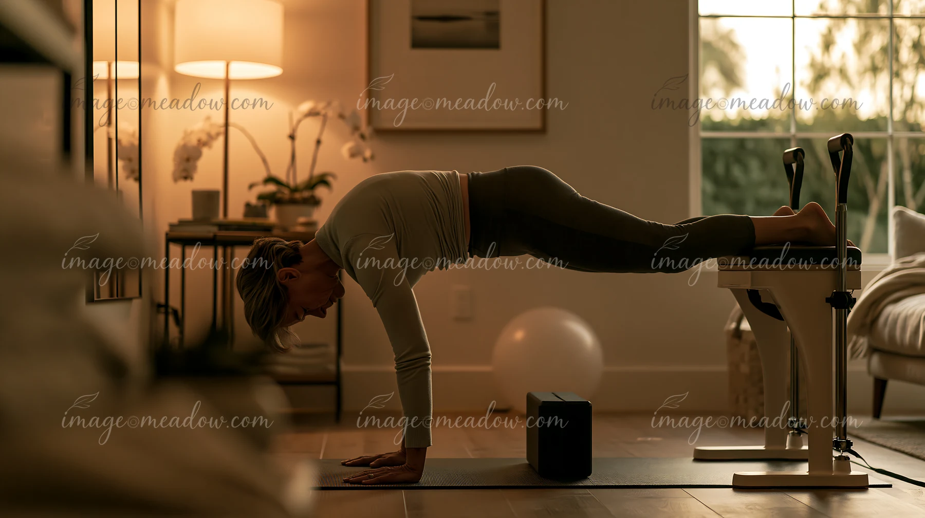 Older woman practices a plank-style Pilates exercise on a mat beside a reformer in a warmly lit home.