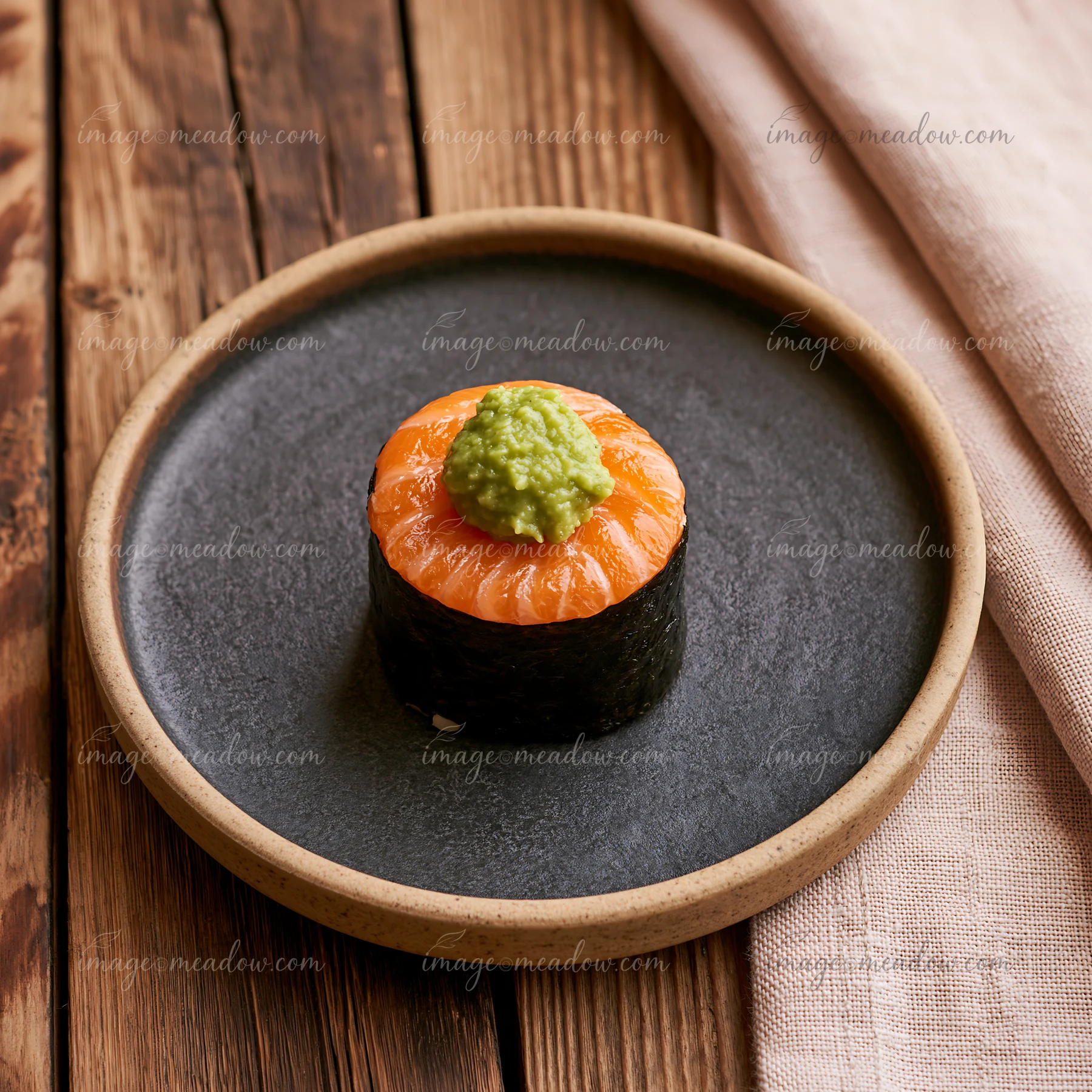One salmon maki piece with wasabi sits on a dark plate placed on a wooden table.