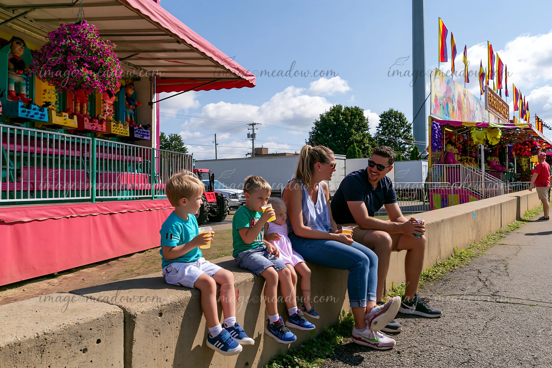 Parents and three children sit beside colorful fairground game stalls while enjoying drinks on a sunny day.