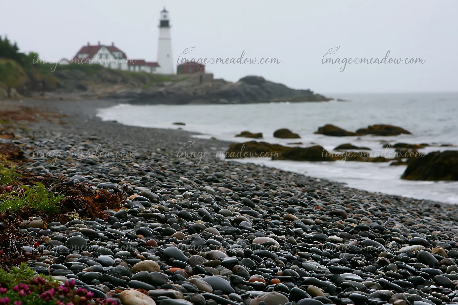 Pebbles cover the beach while waves gently touch the shore near a lighthouse.