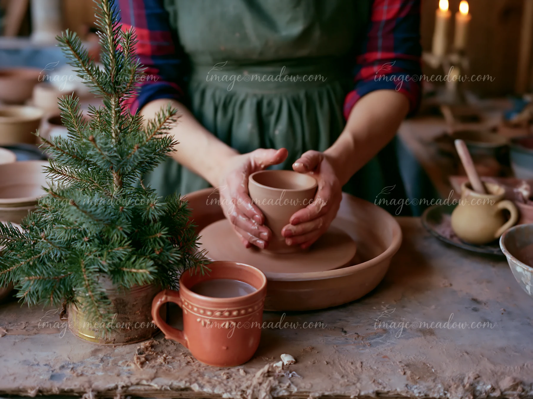 Person holds a ceramic mug beside a small evergreen tree on a rustic table with candles and pottery.