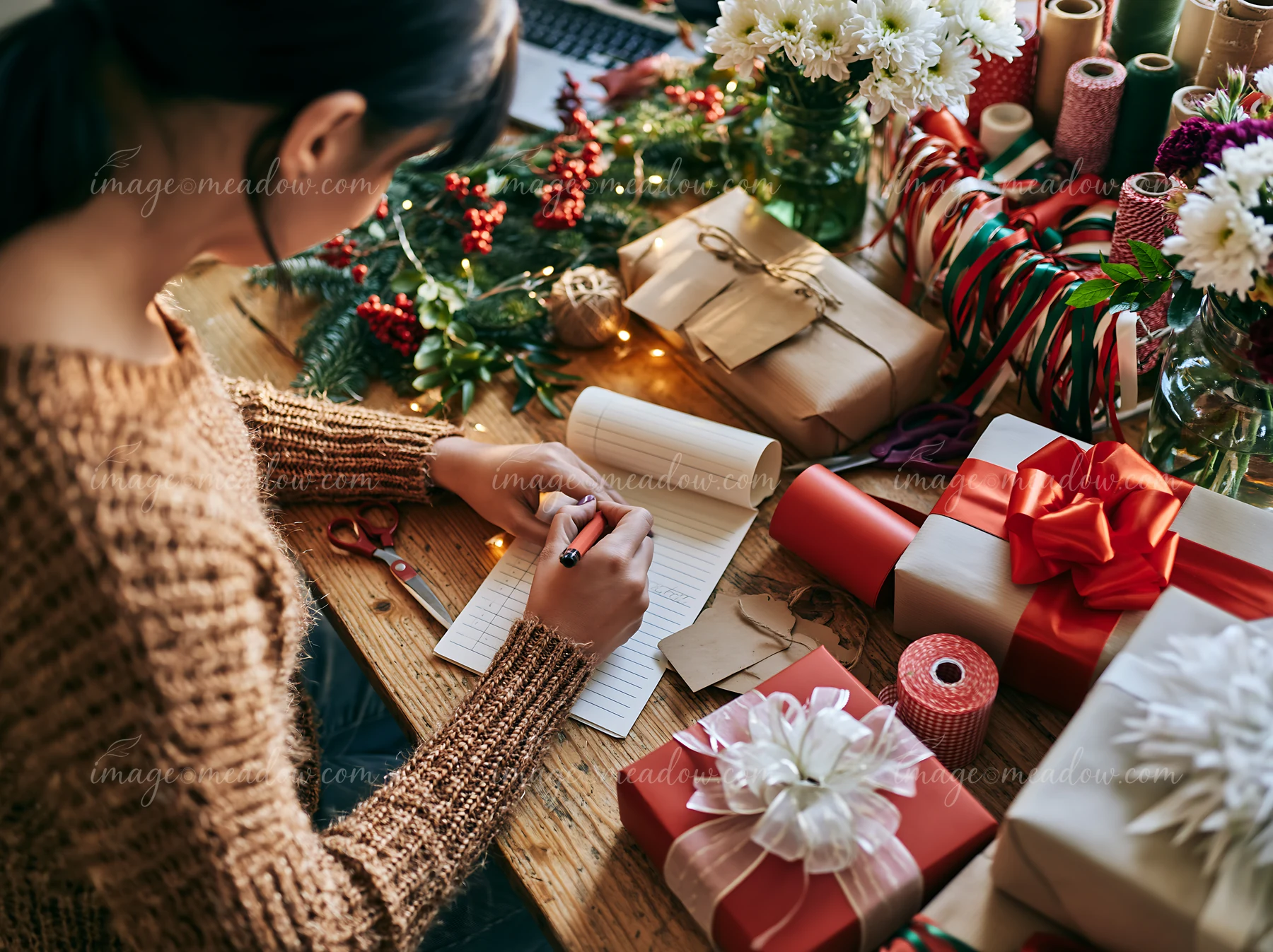 Person writes a gift list at a wooden desk surrounded by wrapped presents, flowers, ribbon, and festive greenery.