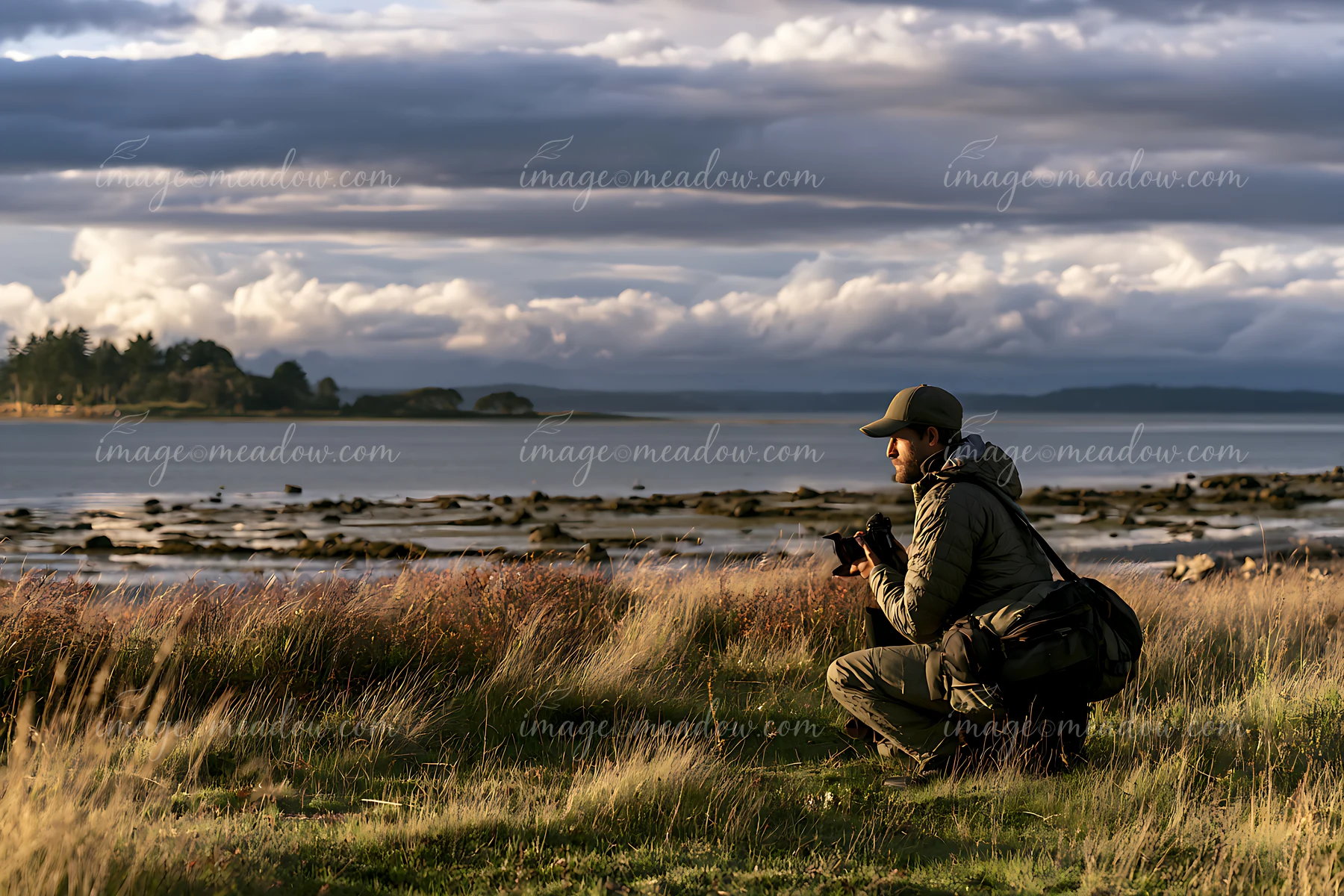 Photographer kneels in a seaside meadow beside a rocky shore beneath dramatic clouds.