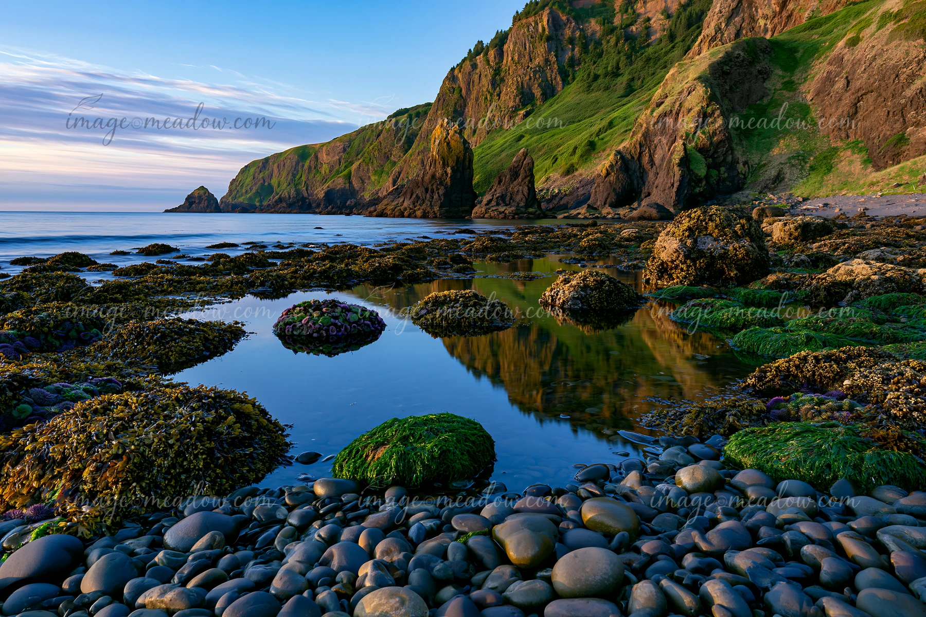 Rocks and pebbles are visible along the coastline beneath dramatic cliffs at low tide, with reflections in calm water.
