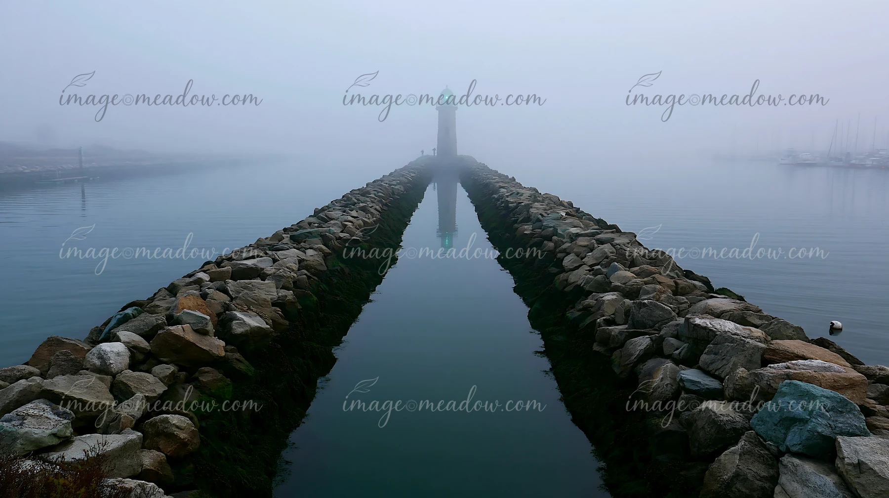 Rocky harbor breakwater leads through calm foggy water toward a distant lighthouse.