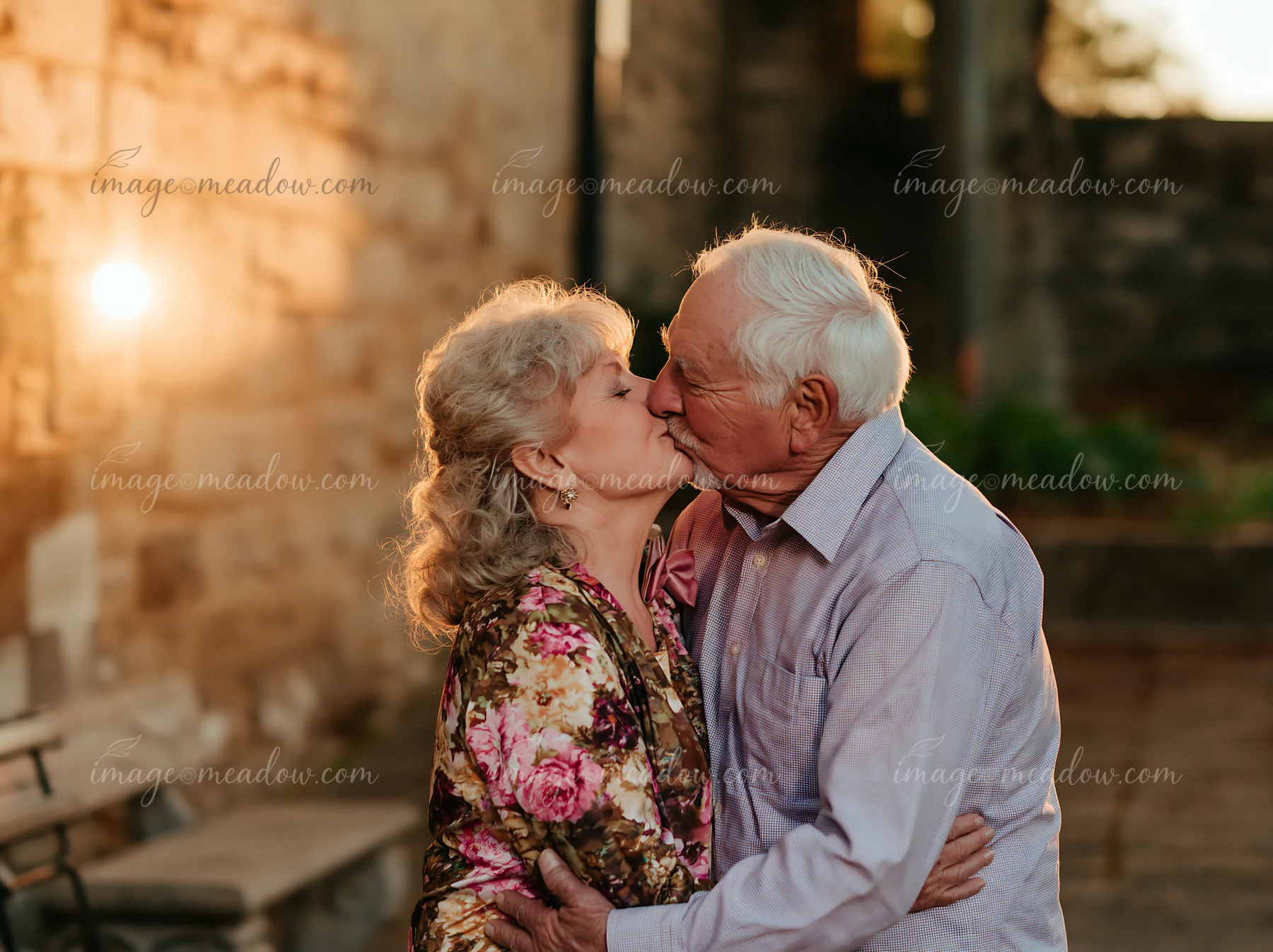 Senior couple kissing outdoors at sunset, warmly lit beside a stone wall with open copy space to the left.