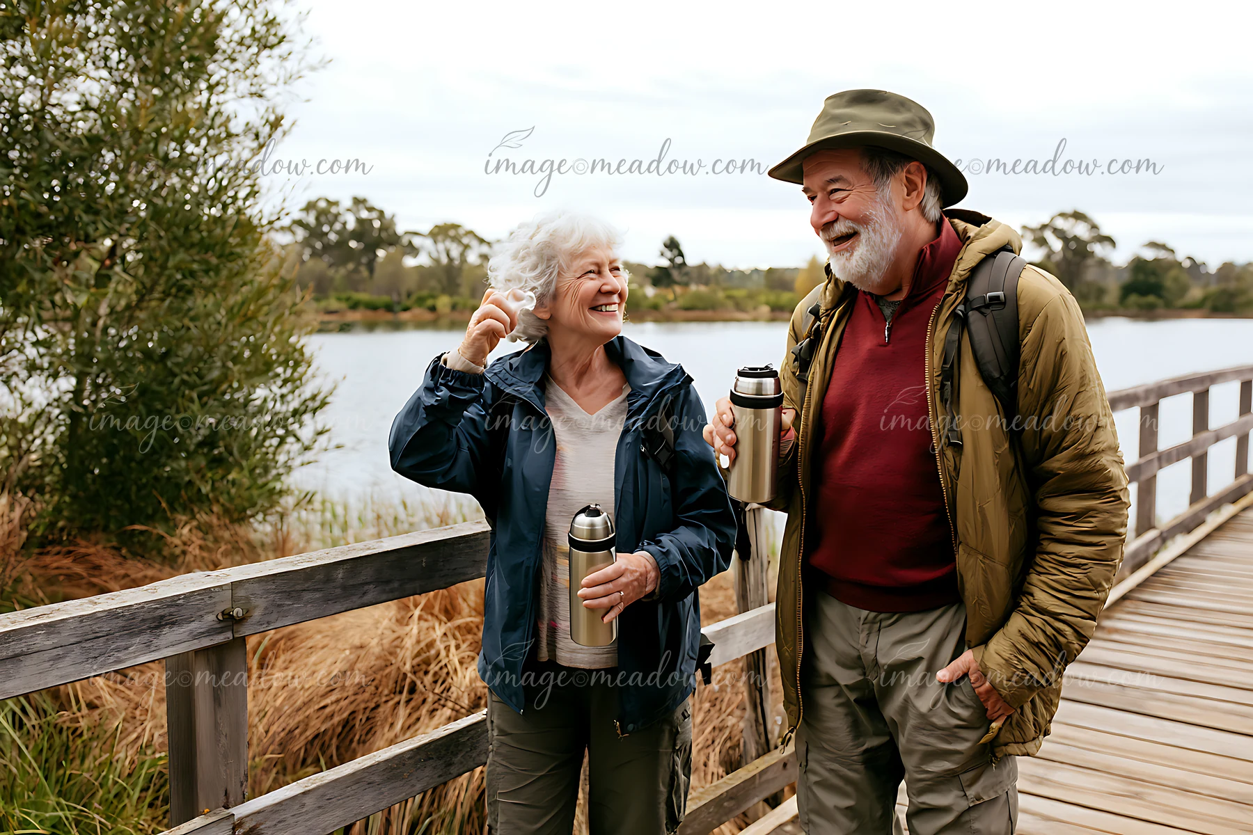 Smiling older couple walks across a wooden footbridge beside a calm lake, carrying backpacks and insulated travel mugs.