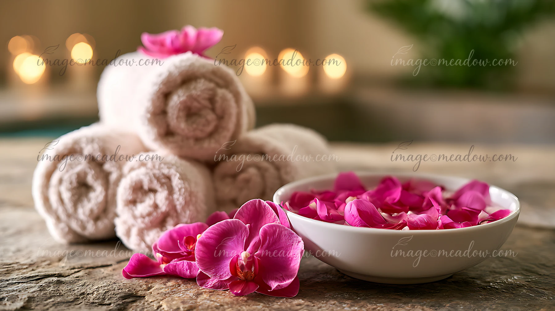 Soft towels are stacked on a stone surface next to a bowl filled with pink flower petals.