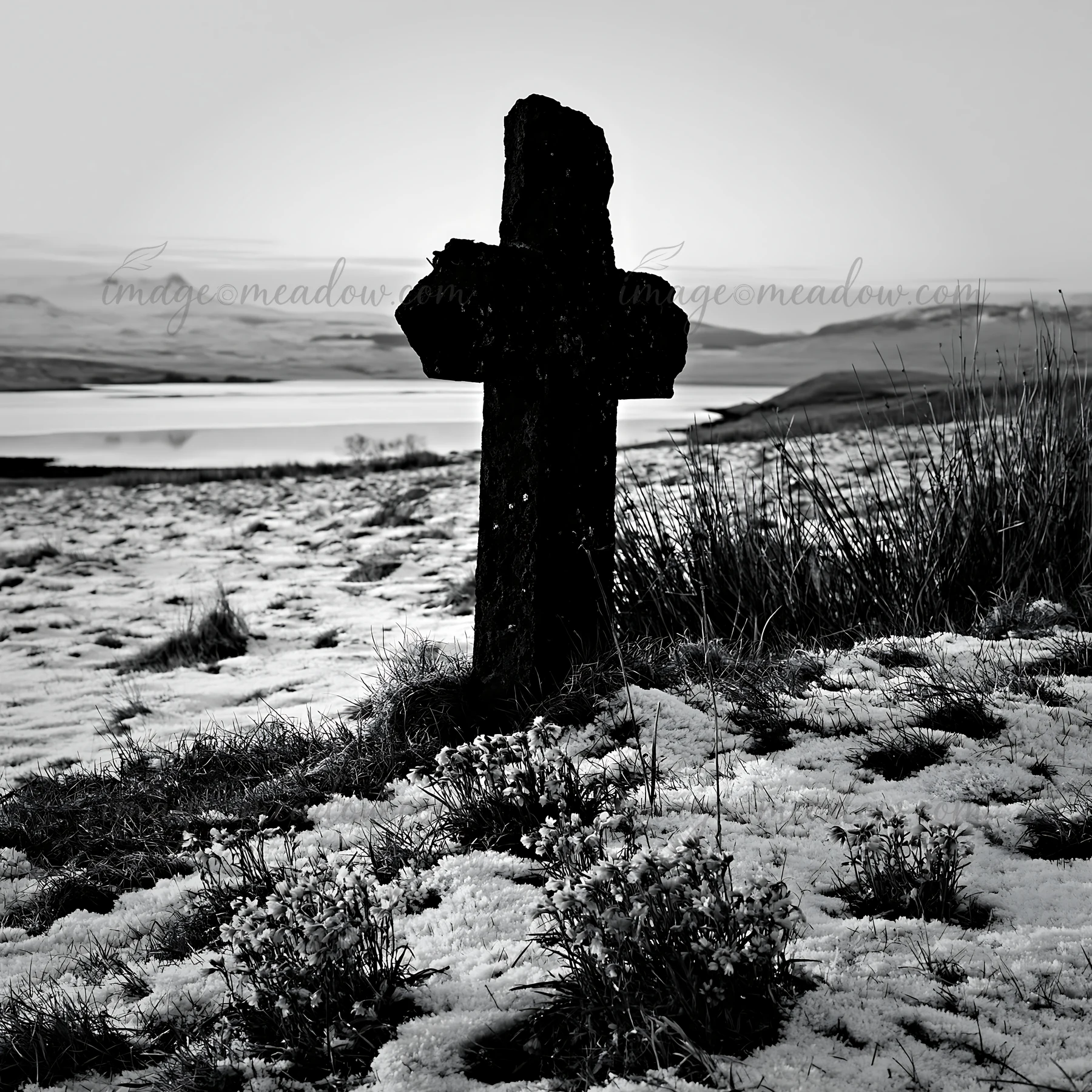 A stone Christian cross stands in a snowy meadow with patches of grass. The landscape features a quiet body of water in the background and distant hills. The se