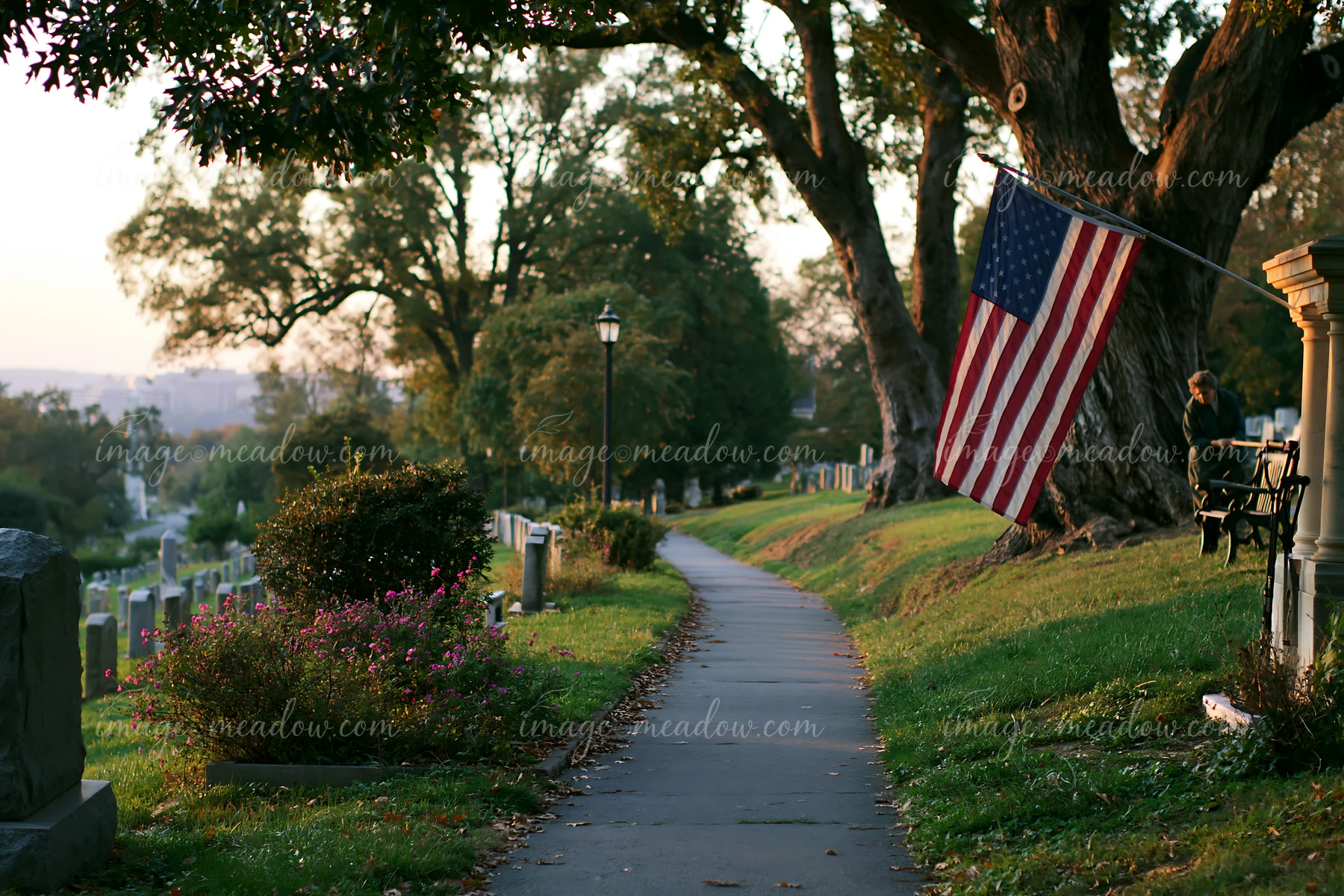 The American flag at Arlington Cemetery is seen along a quiet path lined with gravestones and trees during the golden hour of sunset.