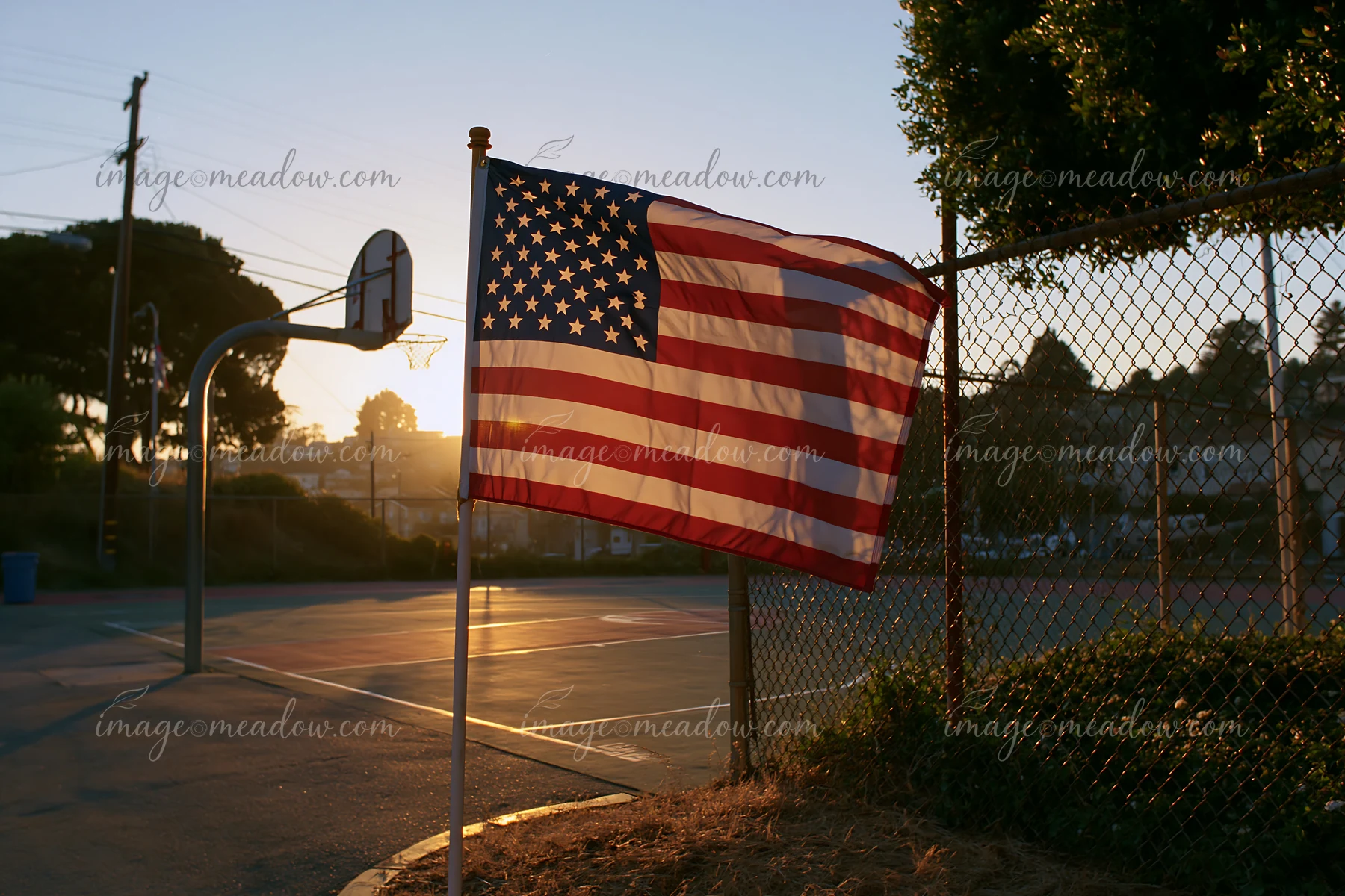 The American flag waving in gentle breeze is displayed on a flagpole near a basketball court at sunset.