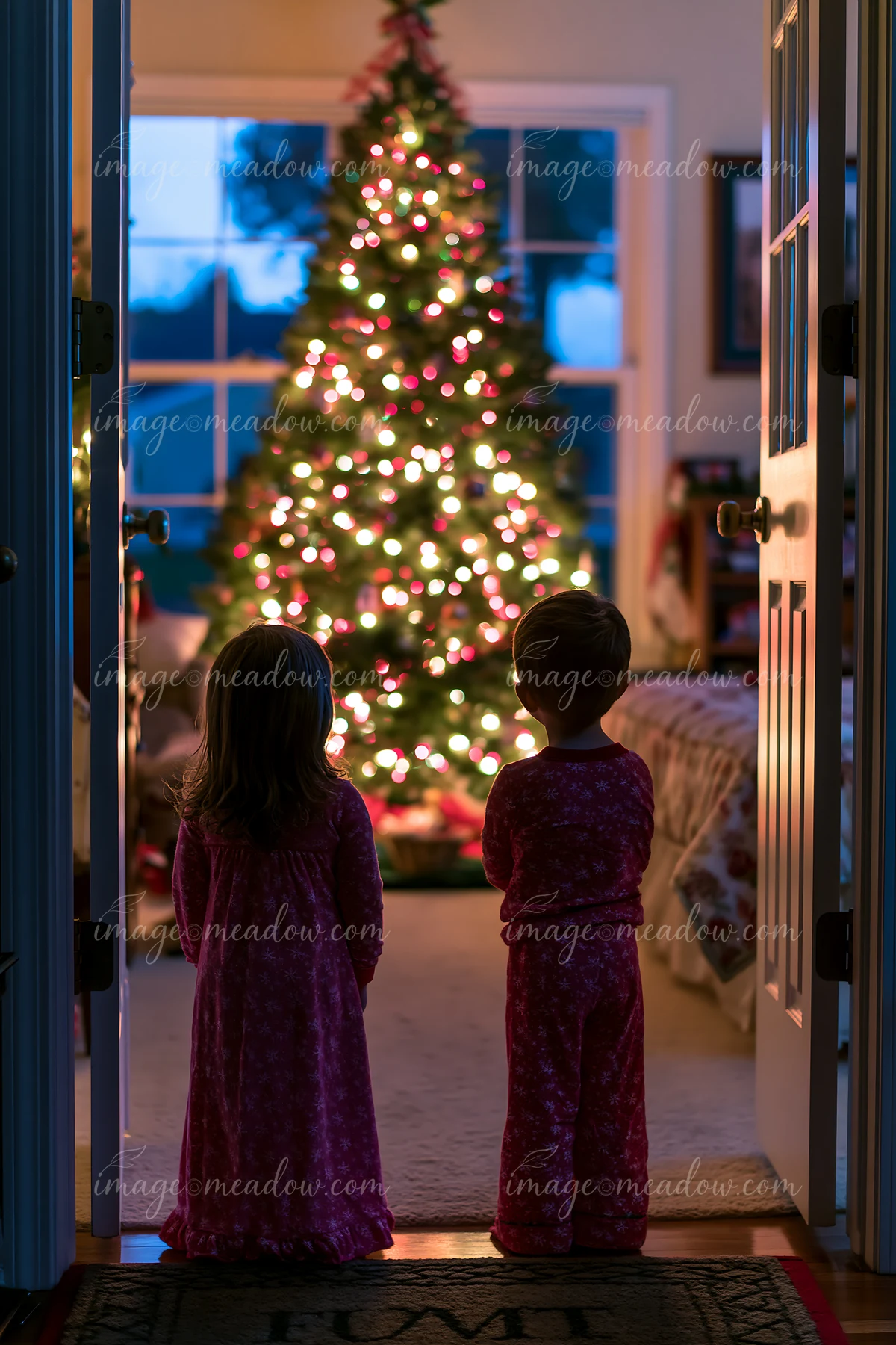 Two children wearing matching pajamas stand in a doorway, admiring a brightly lit Christmas tree in a cozy living room in the evening.