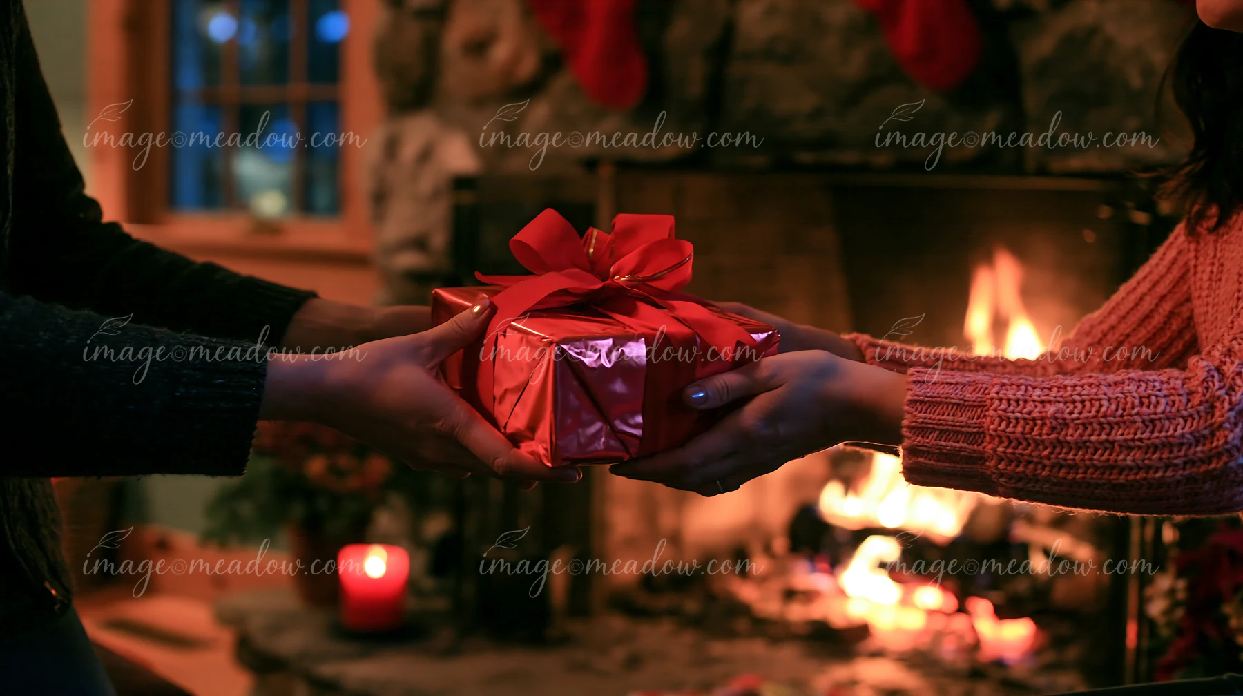 Two hands are exchanging a wrapped gift near a warm fireplace during a holiday gathering.