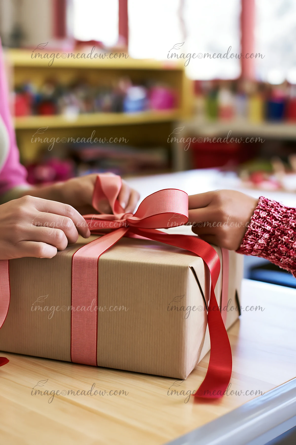 Two hands are tying a pink ribbon around a brown gift box on a table in a classroom.