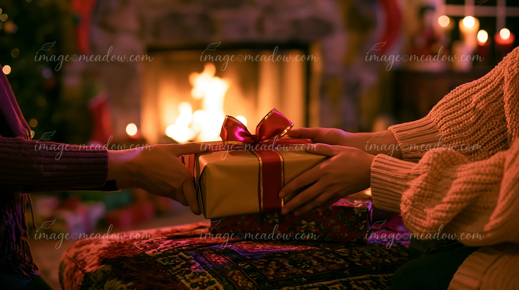 Two pairs of hands exchange a red-ribboned gift in a warmly lit room beside a fireplace.