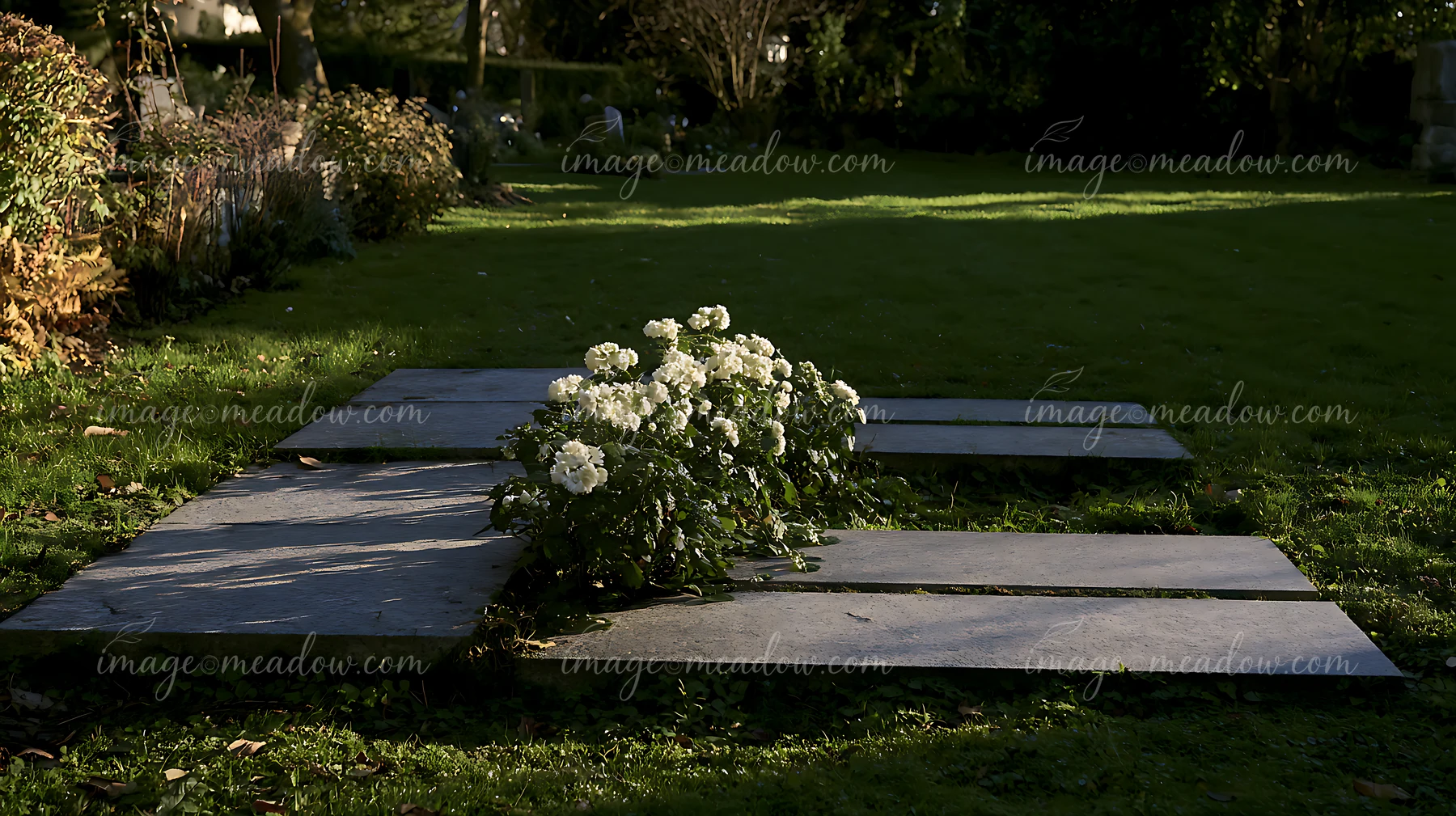 White flowers decorate a flat grave among stone memorials and green lawn in a shaded cemetery.