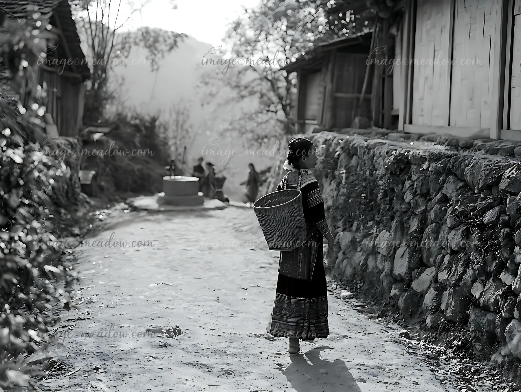Woman in traditional dress carries a large basket along a dirt path beside rustic homes.