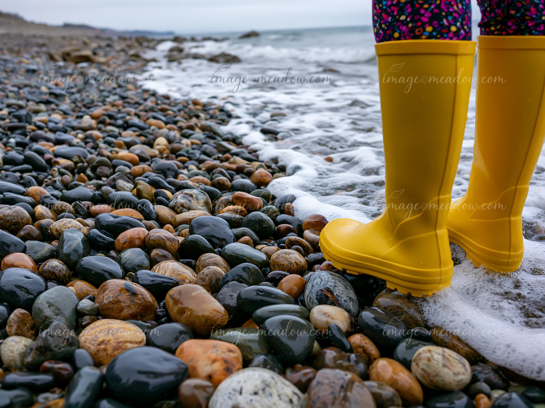 Yellow patterned rain boots stand on wet pebbles beside foamy waves on an overcast coastline beach.