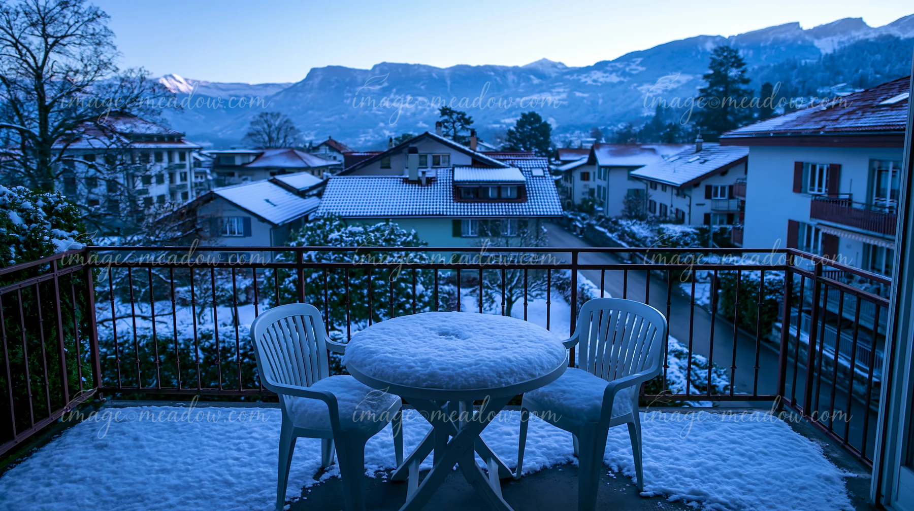 A balcony with two chairs and a round table covered in snow offers a view of Swiss mountain scenery with houses and trees in winter.
