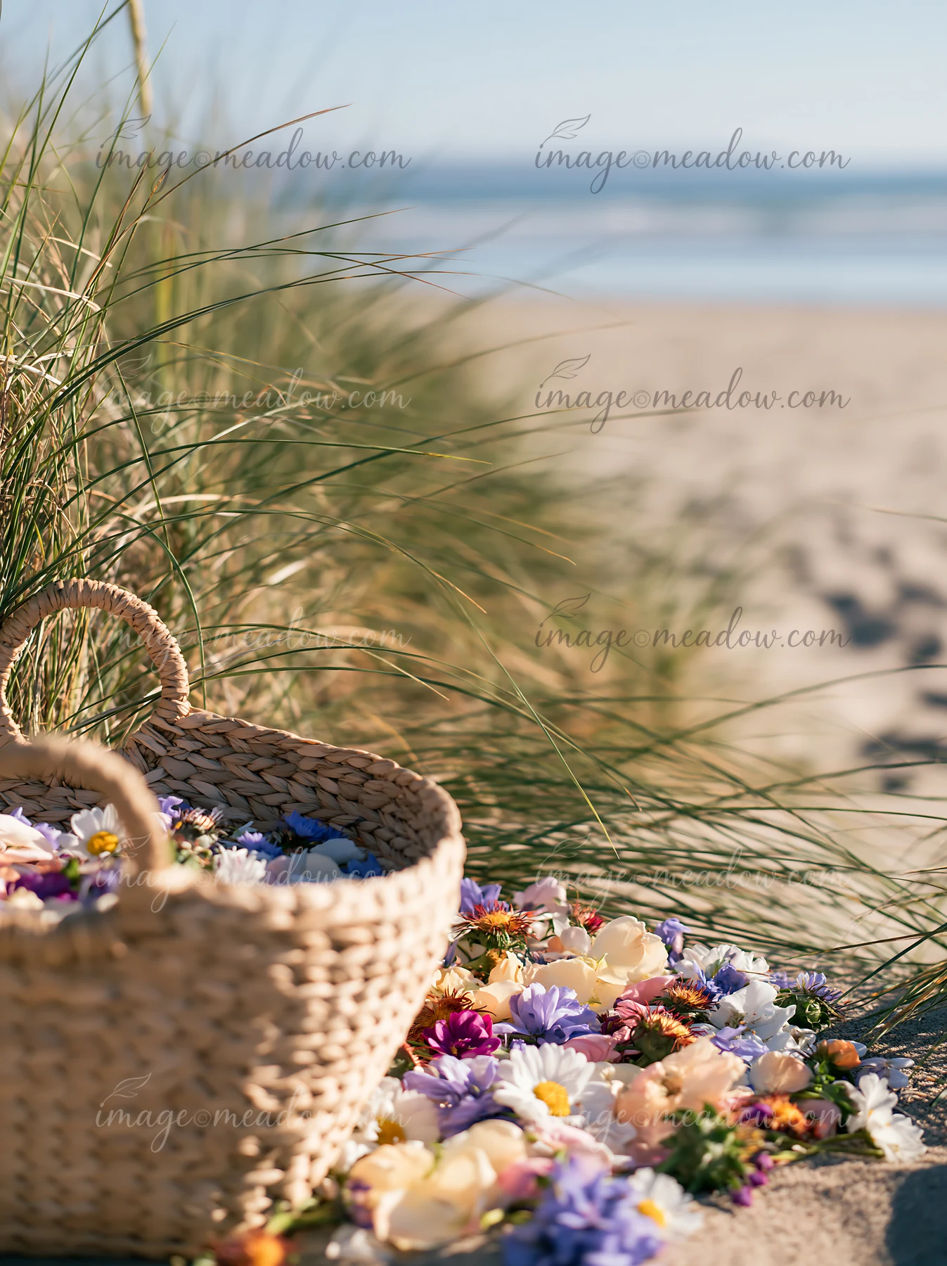 A basket filled with wildflowers lies on the sand near the ocean, surrounded by beach grass.