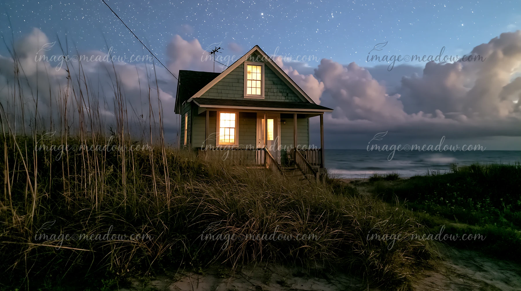 A beach house stands on the shoreline, lit from within, surrounded by tall grass and the ocean.