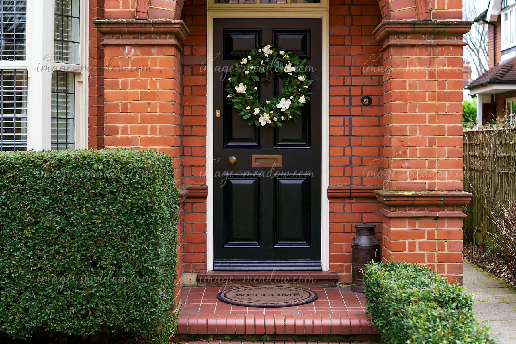 A black front door features a green wreath and a welcome doormat at a brick home entrance.