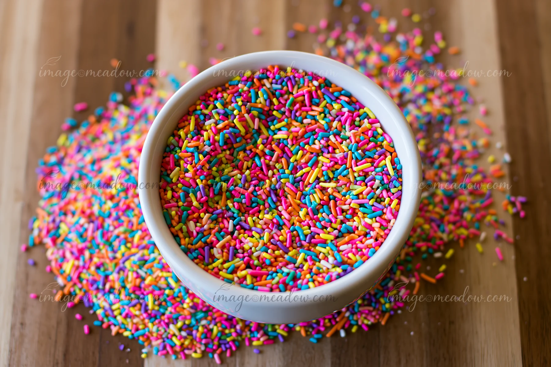 A bowl filled with colorful rainbow sprinkles is placed on a wooden table, surrounded by sprinkles arranged in a swirl pattern.