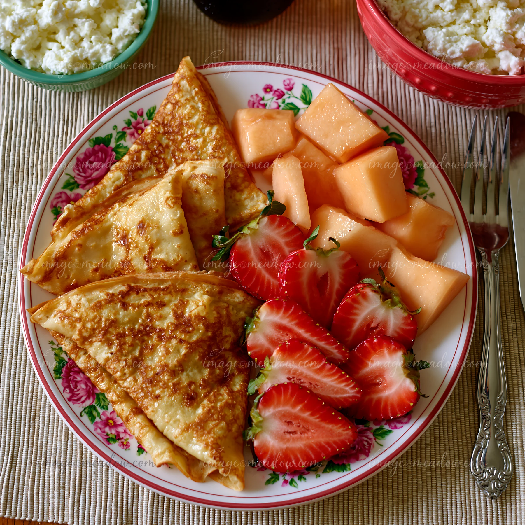 A breakfast plate features sweet crepes alongside fresh strawberries and melon cubes.