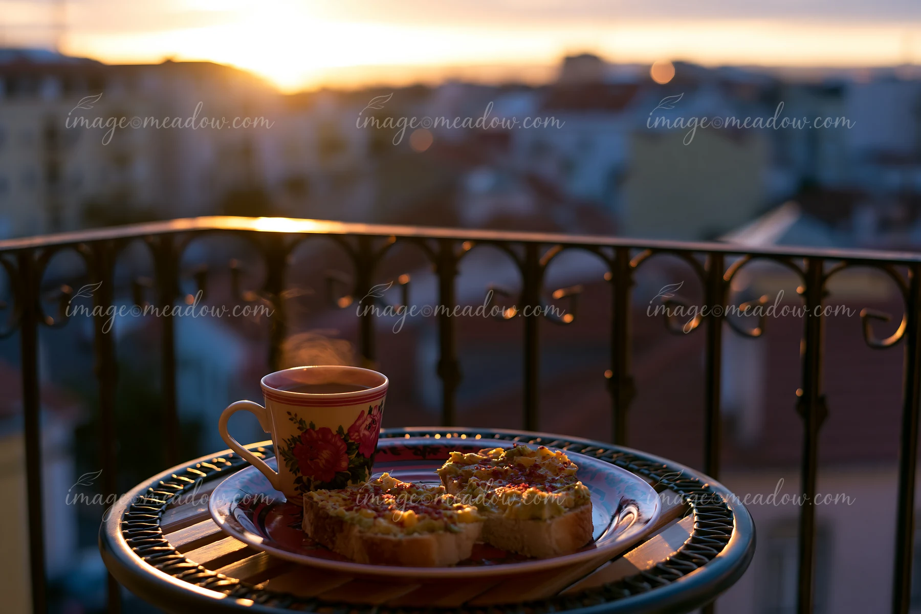 A breakfast plate with avocado toast and a cup of coffee sits on a balcony table as the sun rises over the city skyline.