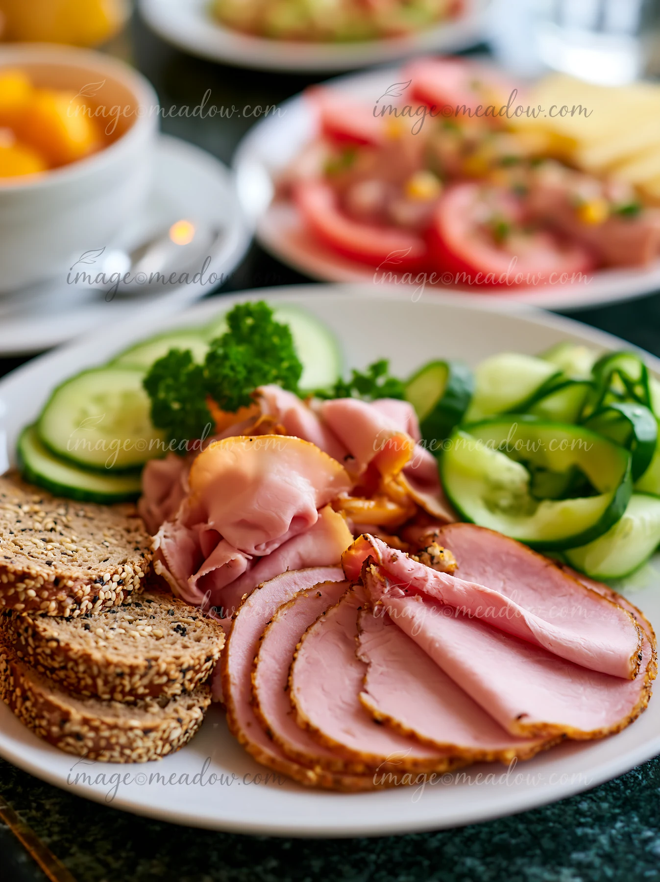 A breakfast plate with cold cuts includes slices of ham, turkey, and bread alongside cucumber, parsley, and a side of yogurt.