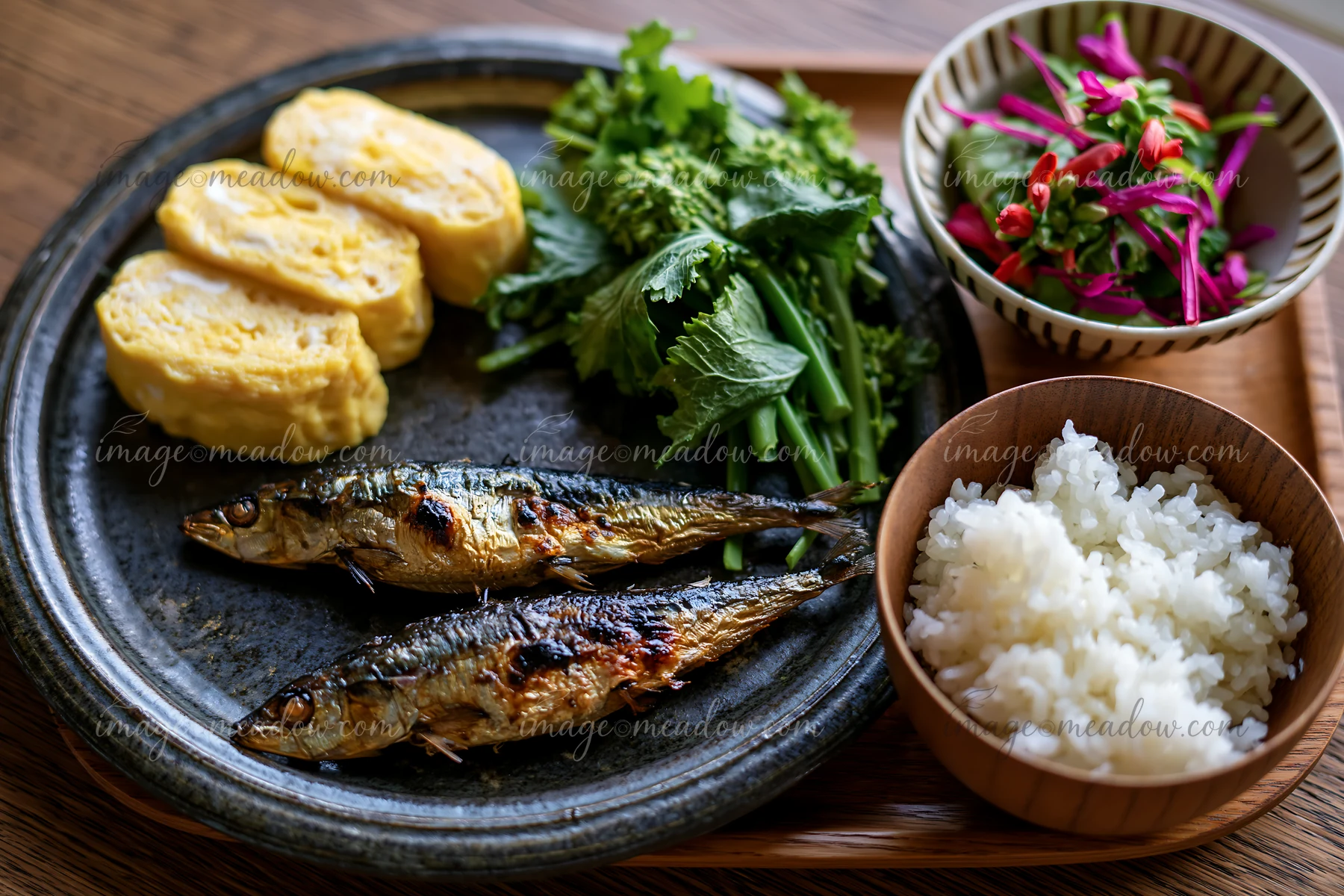 A breakfast plate with Japanese breakfast features grilled fish, rolled omelette, greens, and rice.