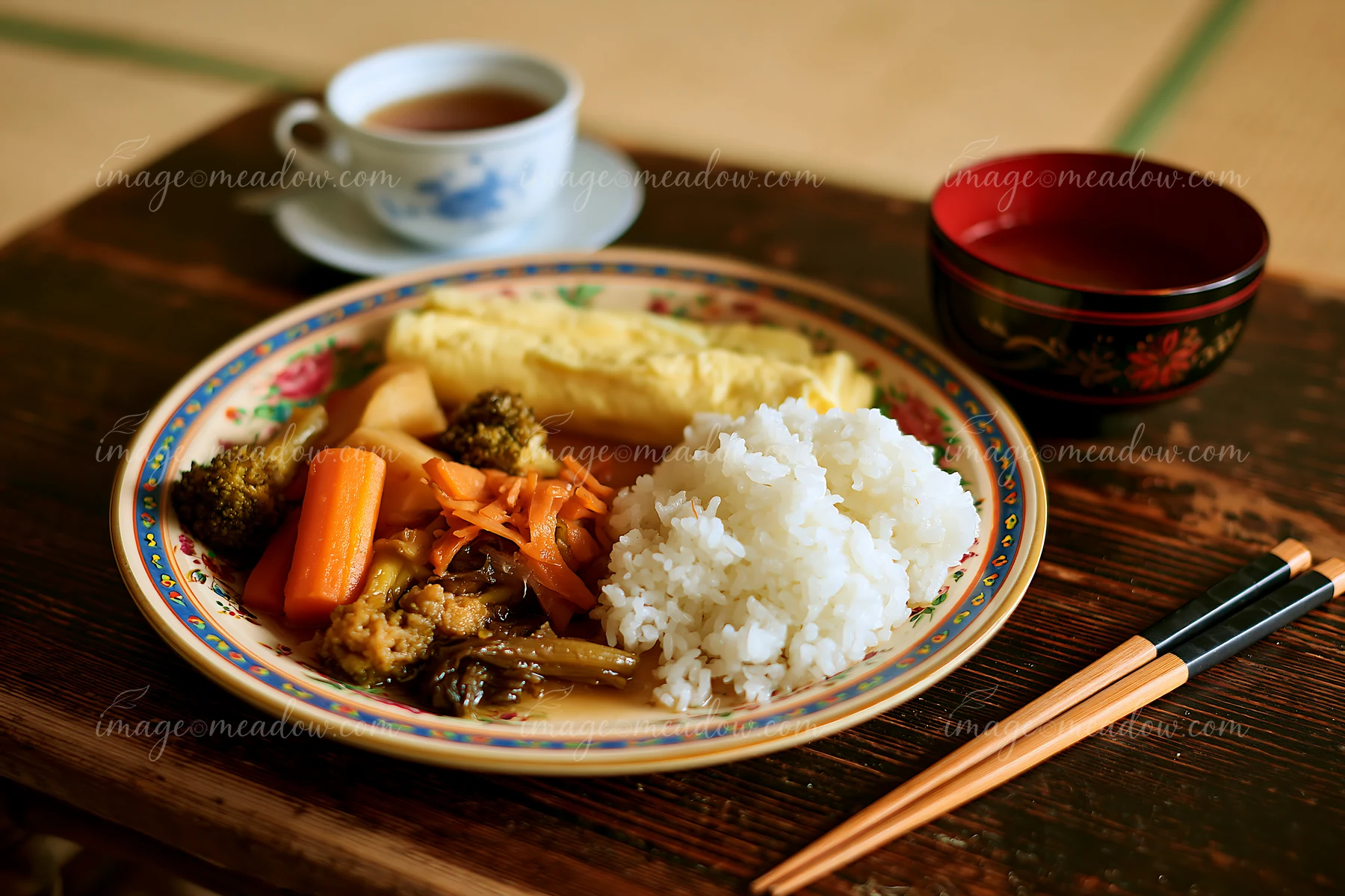 A breakfast plate with Japanese breakfast includes rice, vegetables, and an omelet alongside a cup of tea, arranged on a wooden table in a traditional setting.