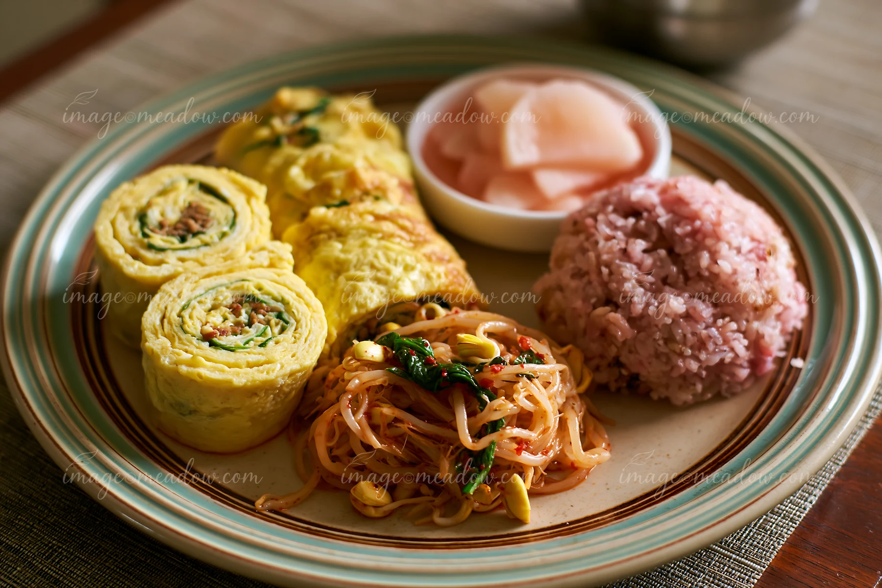 A breakfast plate with Korean breakfast includes rolled omelets, stir-fried noodles, pink rice, and pickled radish.