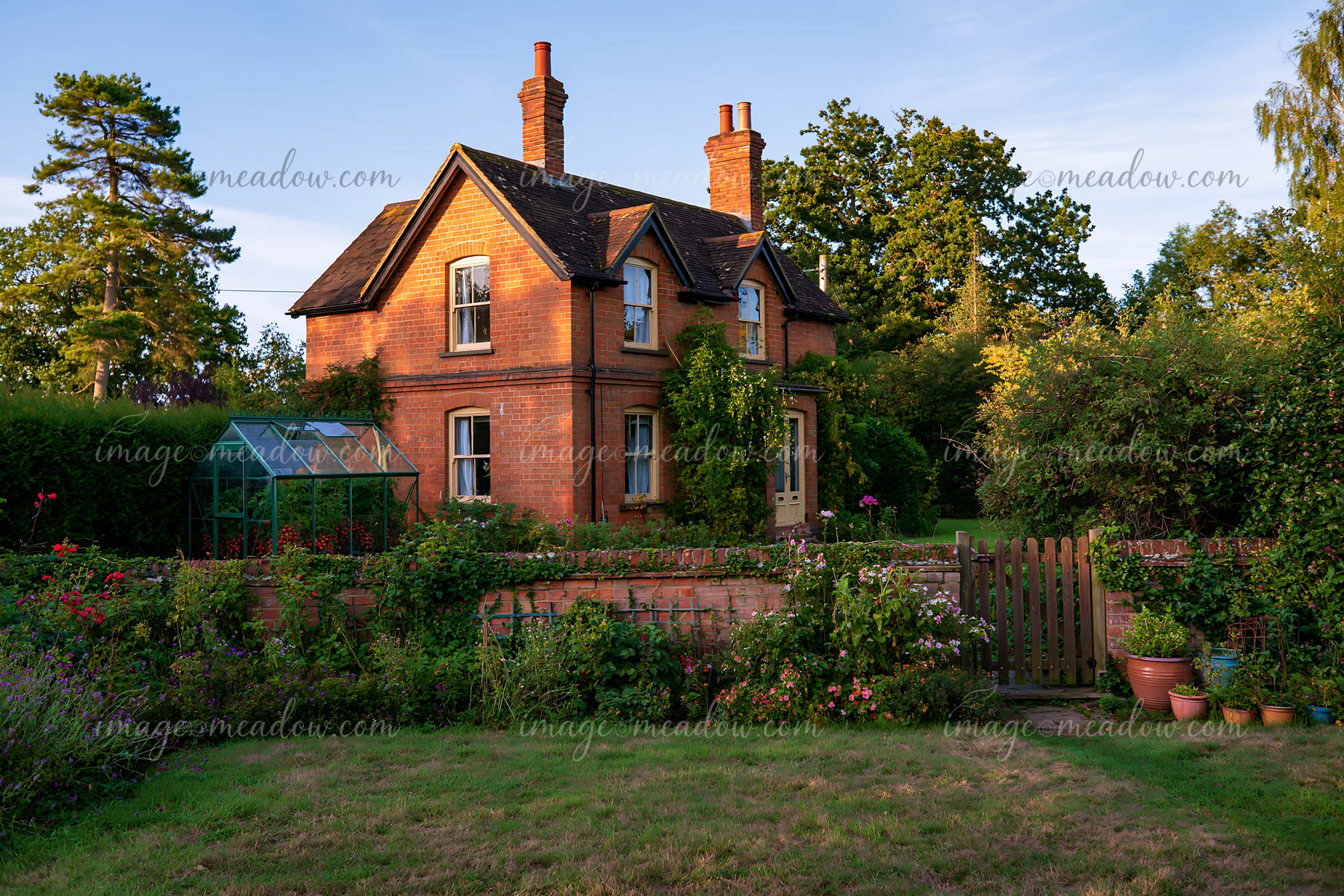 A brick country house with a green garden is bordered by thick hedges.