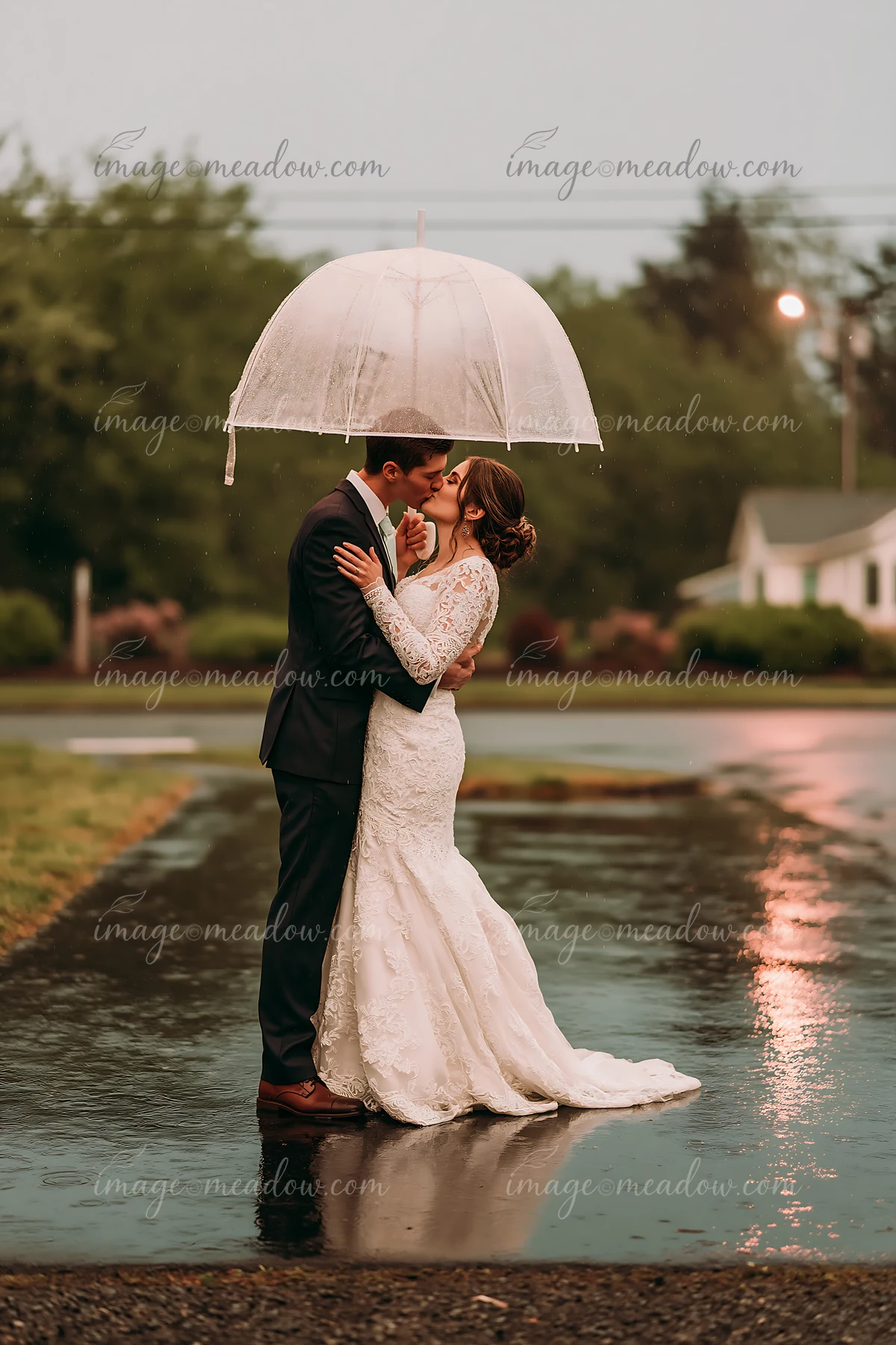 A bride and groom share a kiss under an umbrella in the rain, standing on a wet street after their wedding ceremony.