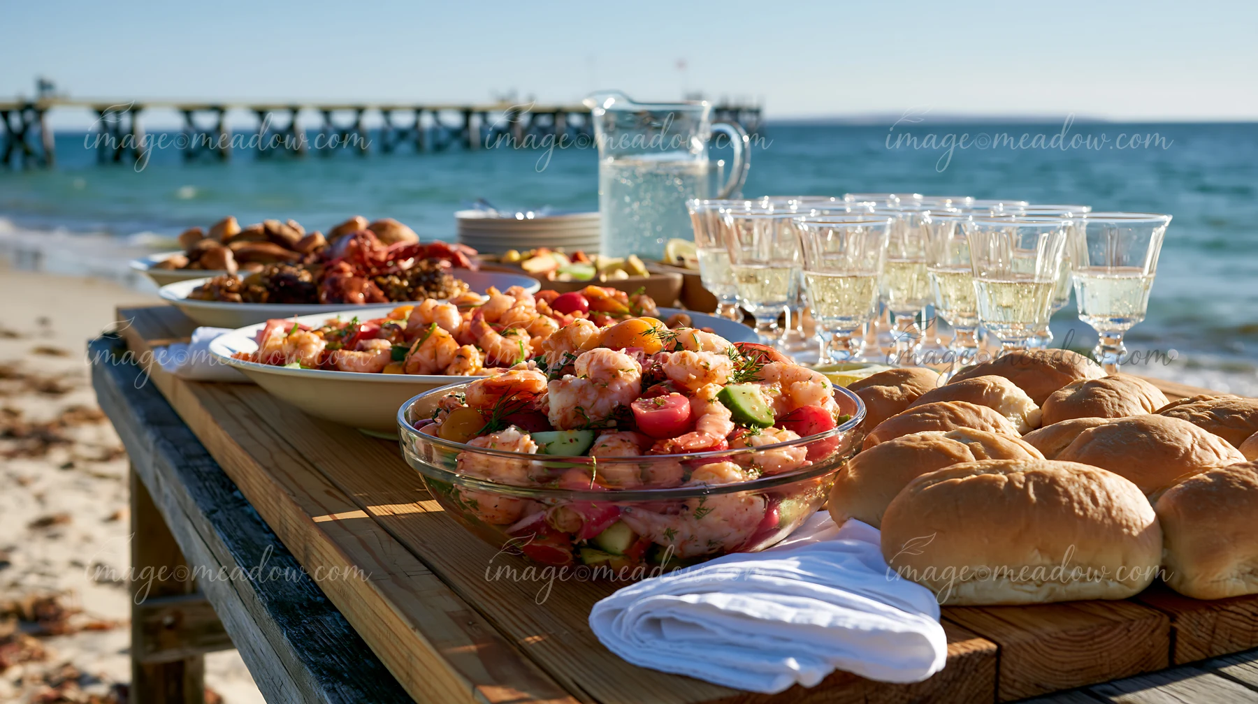 A buffet spread features seafood dishes and beverages on a wooden table by the beach.
