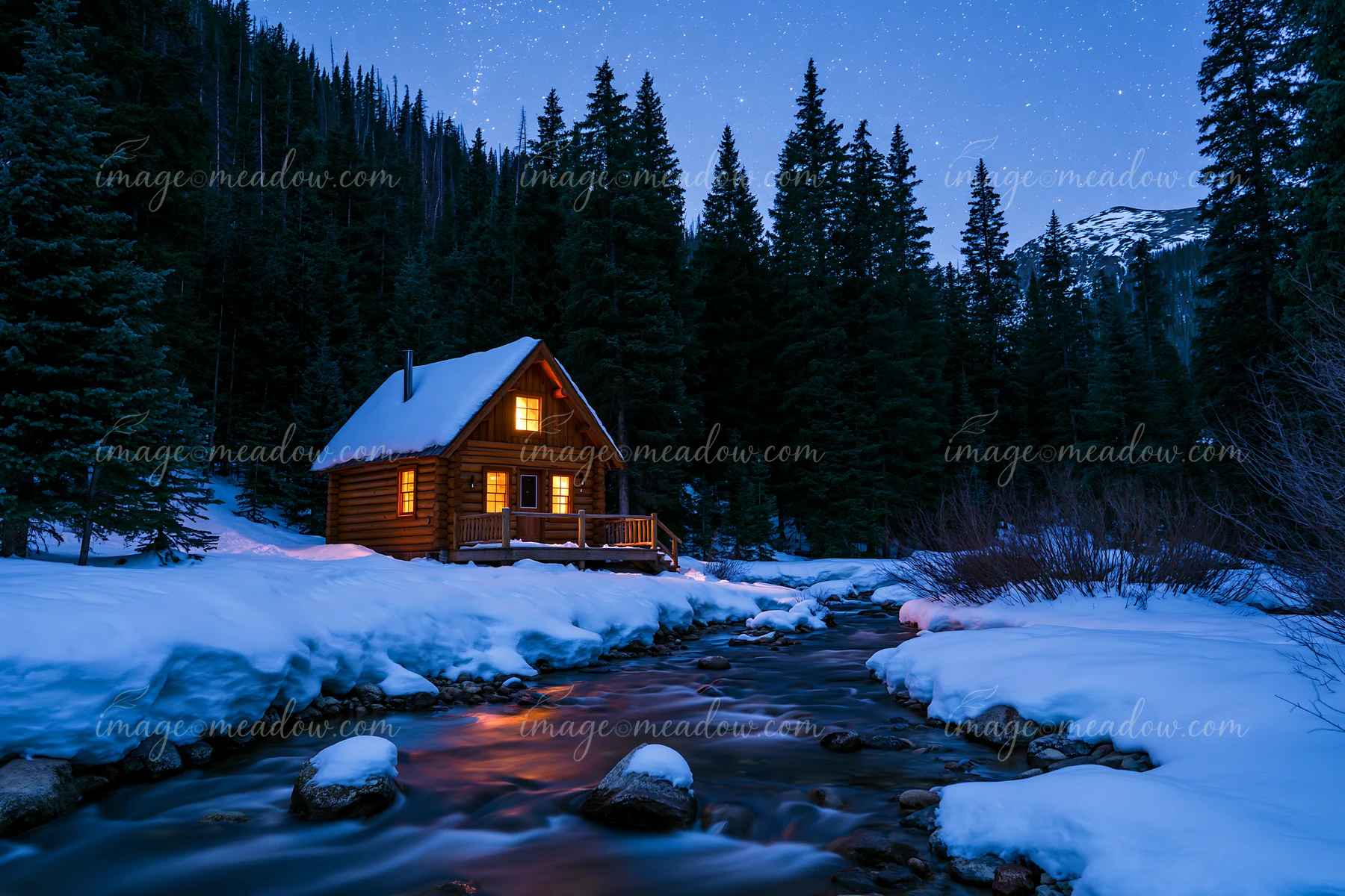 A cabin sits beside a stream, surrounded by snow and tall trees, under a starry sky.