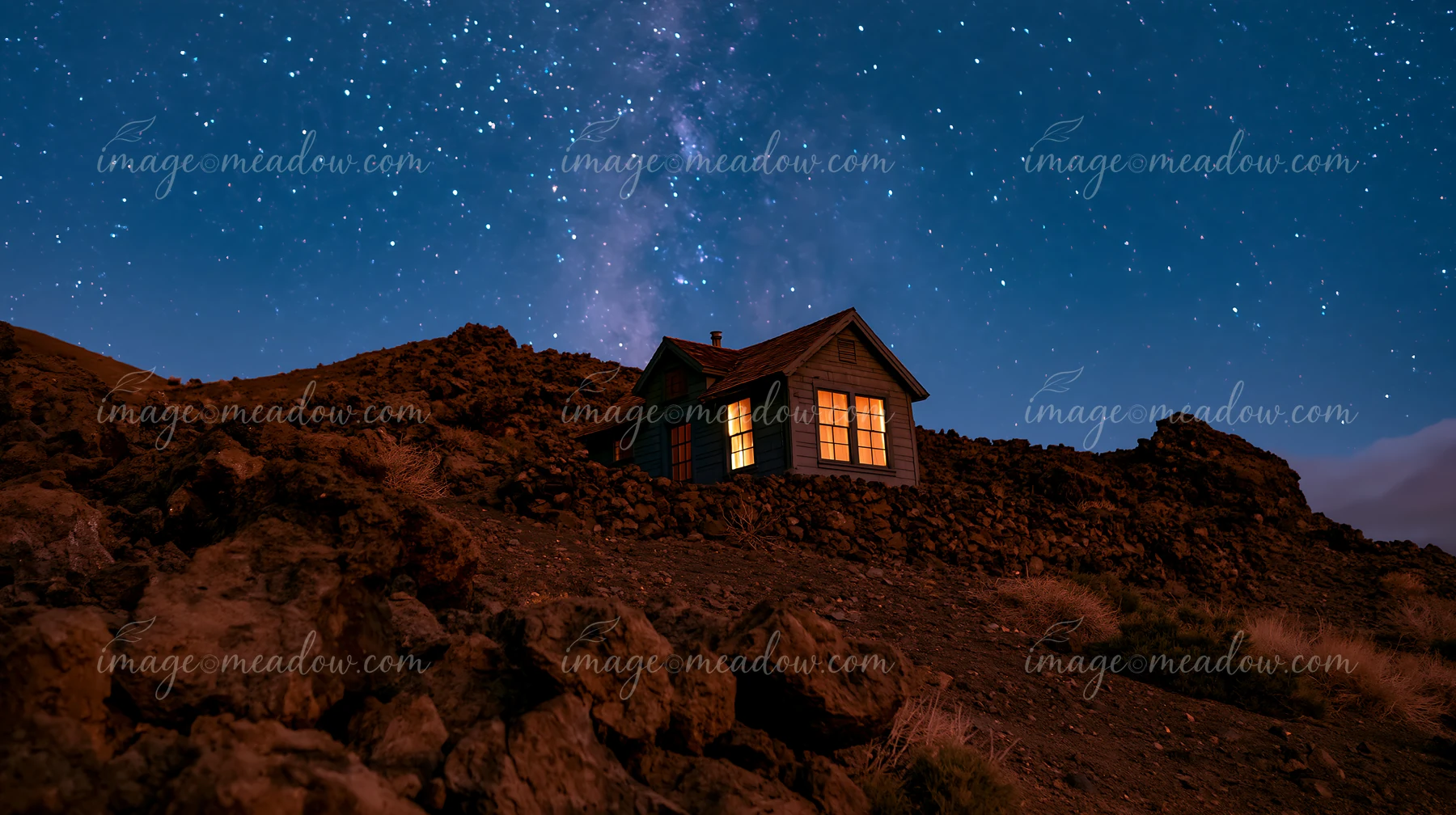 A cabin stands alone on rocky terrain with bright windows, surrounded by a dark night sky filled with stars and the Milky Way.
